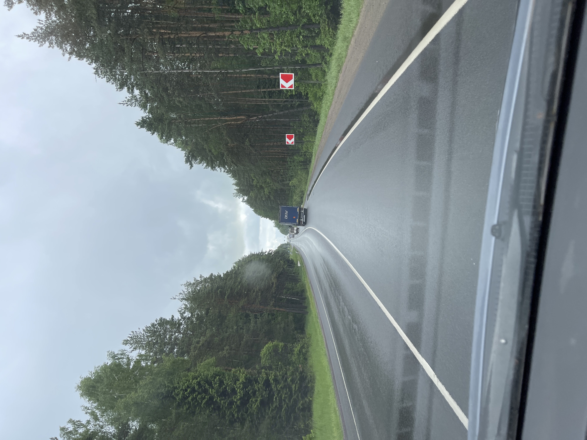 The image depicts a view from inside a vehicle traveling on a wet, asphalt road in Semyonovskoye, Russia. The road is bordered on both sides by dense forest. On the right side of the road are two signs with red chevrons pointing left. A blue truck is visible in the distance, along with other vehicles. The sky is overcast, suggesting rain or a recent rain event. The image is captured from inside the vehicle, evident by a portion of the car's frame visible on the right.