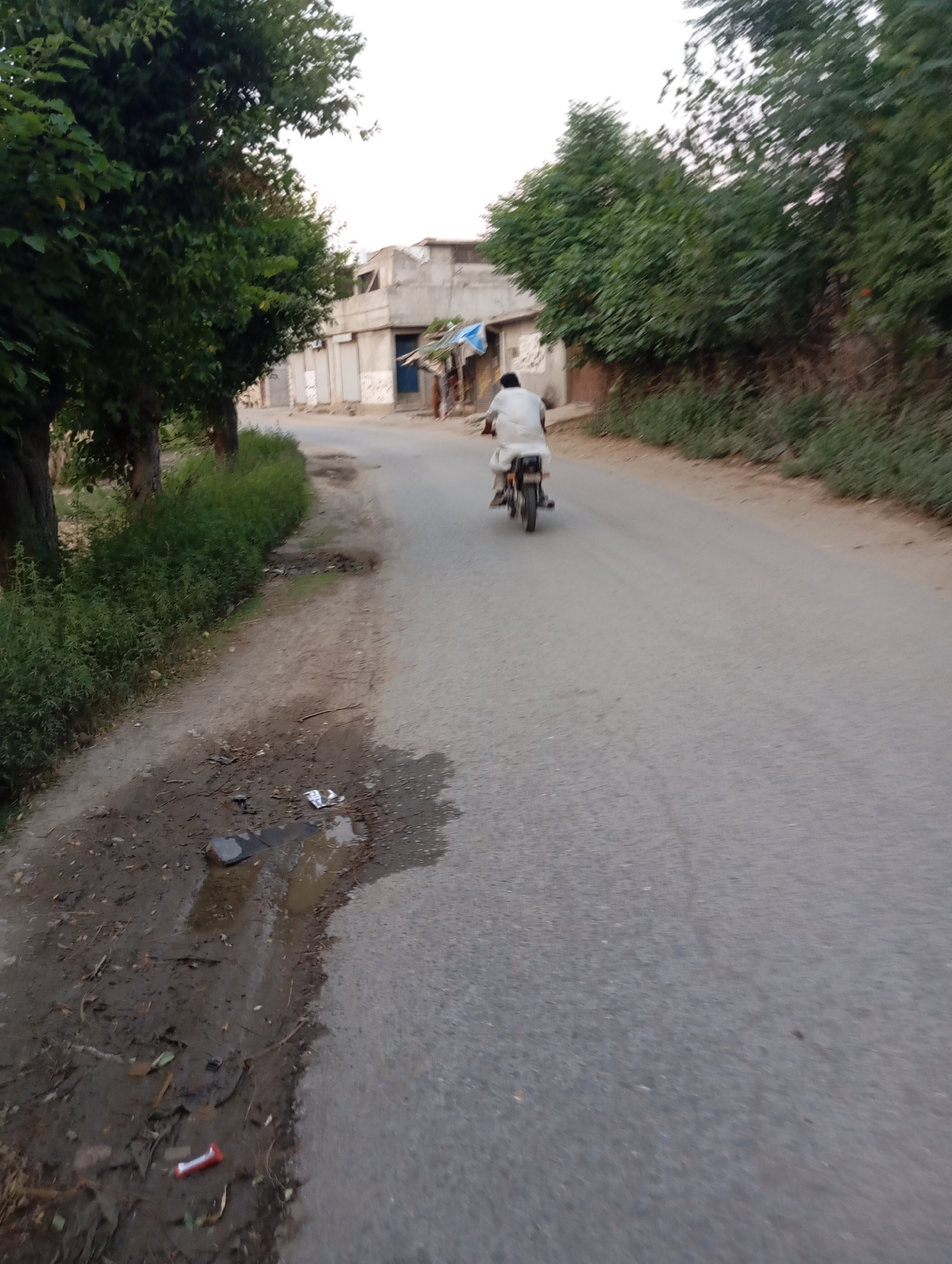 An overhead, slightly tilted, medium shot shows a person riding a motorcycle down a paved road in Lassan, Pakistan. The person is wearing a white outfit. The road is on the right side of the frame, and on the left is a dirt path. There are trees on the far left and in the background on the right. Buildings are visible in the background on the right. The sky is a light gray.