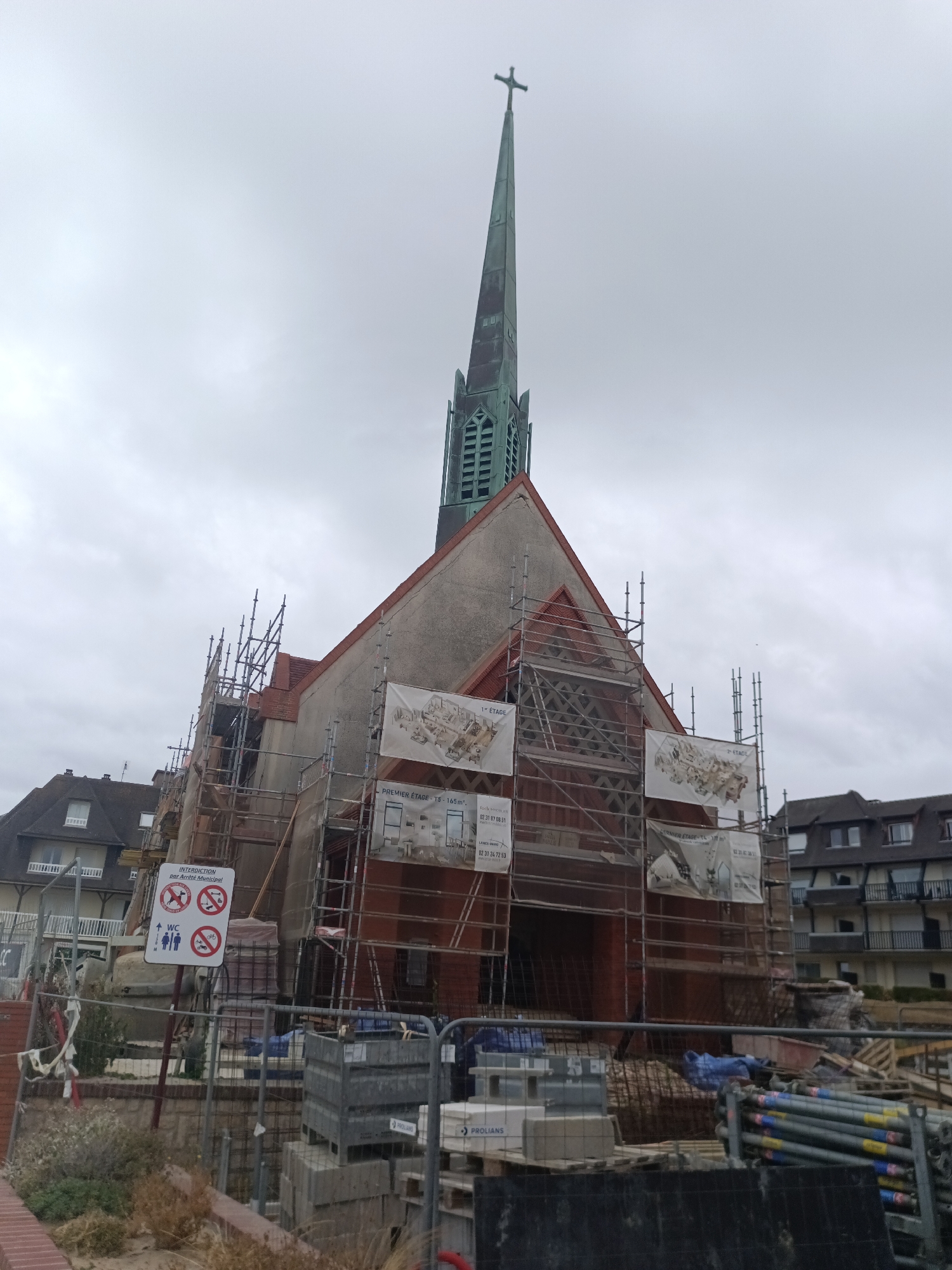 This media file captures an exterior view of a church undergoing renovation. The church features a tall, slender green spire topped with a cross, reaching towards an overcast sky. Scaffolding encircles the building, indicating ongoing construction or repair work. Large banners are displayed on the scaffolding, some featuring architectural drawings and text, including "PREMIER ÉTAGE - 75 - 165 m²".
In the foreground, a red brick wall and fencing delineate a construction site. Various construction materials are visible, including stacks of bricks, metal pipes, and blue tarpaulins. A white sign with red lettering reads "INTERDICTION par Arrêté Municipal" (Prohibition by Municipal Decree) and displays several prohibition symbols: a dog, a person with a briefcase (possibly representing work), and a bicycle, along with a sign indicating a public restroom.
The scene is set in Houlgate, France, as suggested by the location context. The cloudy weather creates a subdued atmosphere. The overall impression is one of a community project focused on preserving or improving a significant building. The image does not depict any people actively working on the site, but the presence of construction equipment and signage clearly indicates an active renovation event.