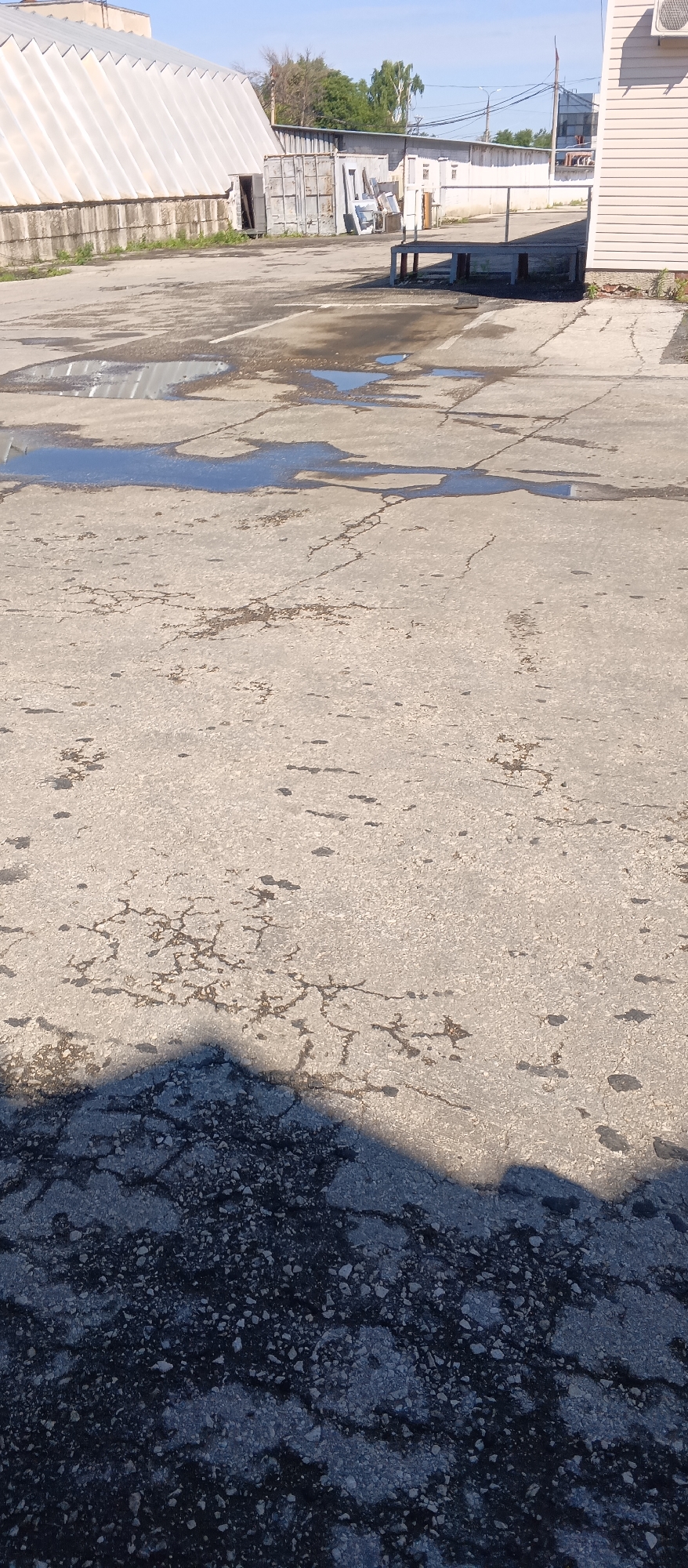 A low-angle, full shot captures a cracked asphalt lot with puddles reflecting a clear blue sky. In the background, a large white industrial building with a corrugated metal roof dominates the left side. To its right, a series of smaller, light-colored buildings and structures are visible, including a metal storage unit and what appears to be a loading dock or platform. The ground in the foreground is a patchwork of dark, rough asphalt and lighter concrete, with several large, irregularly shaped puddles of water scattered across the surface. A dark triangular shadow, likely cast by an unseen object, covers the bottom left portion of the frame. The overall scene suggests an industrial or commercial area, possibly after rainfall.