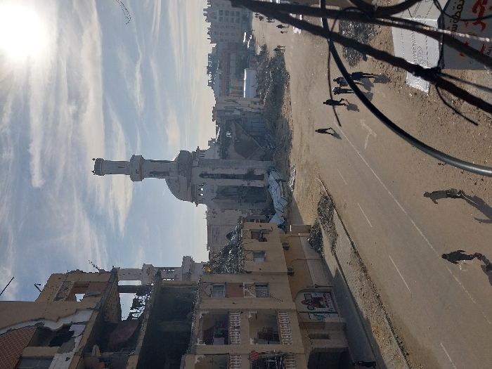 The image shows a scene of devastation, likely the aftermath of conflict. A partially destroyed building stands prominently, with a minaret and dome structure appearing relatively intact amidst the rubble. A road runs through the scene with a few people walking on it. Debris and damage are visible across the cityscape, suggesting widespread destruction. The sky is bright with sunlight and light clouds. Overhead powerlines and a sign are also visible.