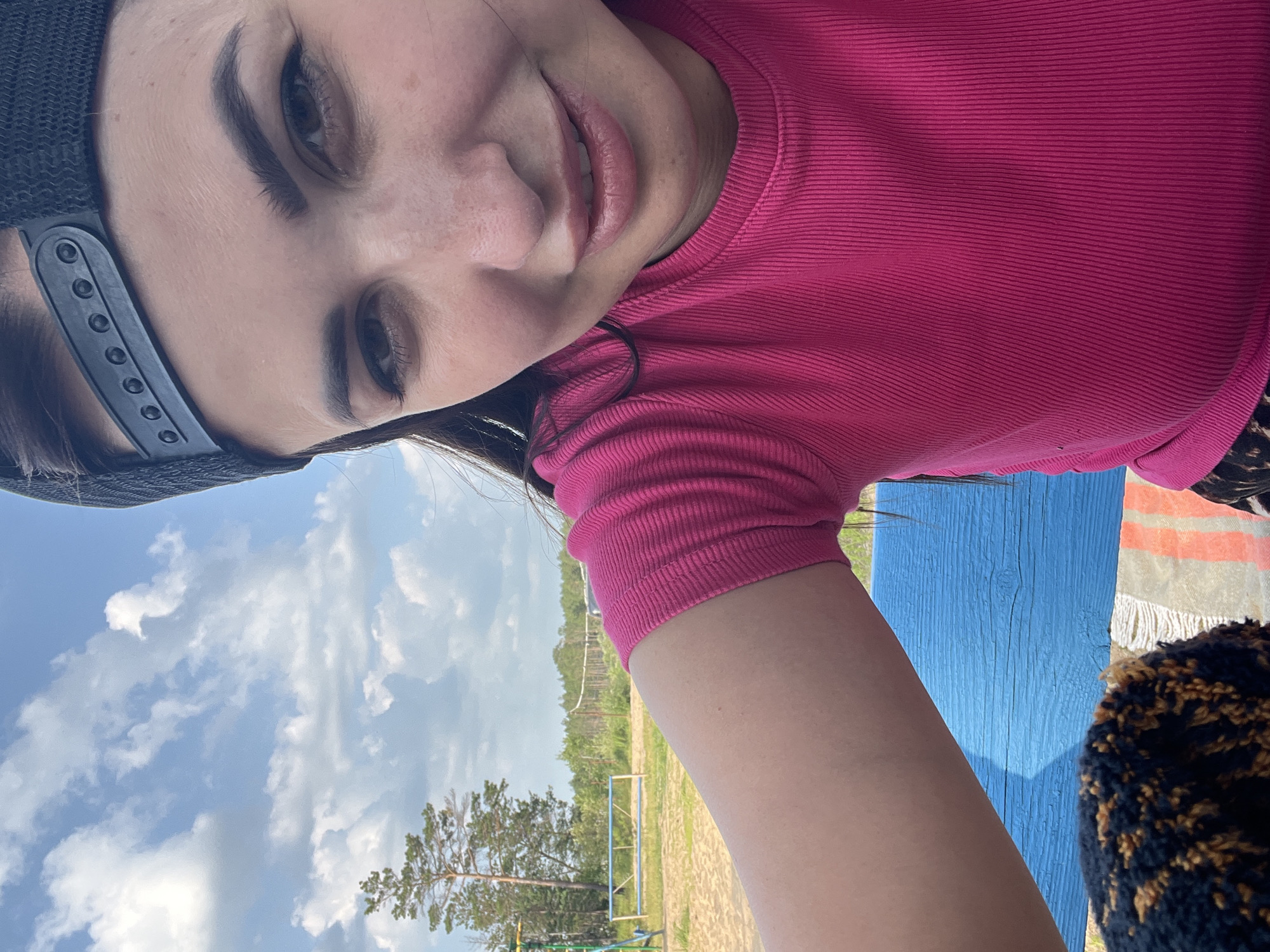 In a close-up, selfie-style shot, a young woman with dark hair, wearing a pink ribbed t-shirt and a black baseball cap, is captured outdoors. Her gaze is directed towards the camera, and she has a subtle smile. The background reveals a bright, partly cloudy sky above a tree line and a sandy area, suggesting an outdoor recreational space. A blue wooden bench is partially visible in the foreground. The scene appears to be in a park or a similar outdoor setting.