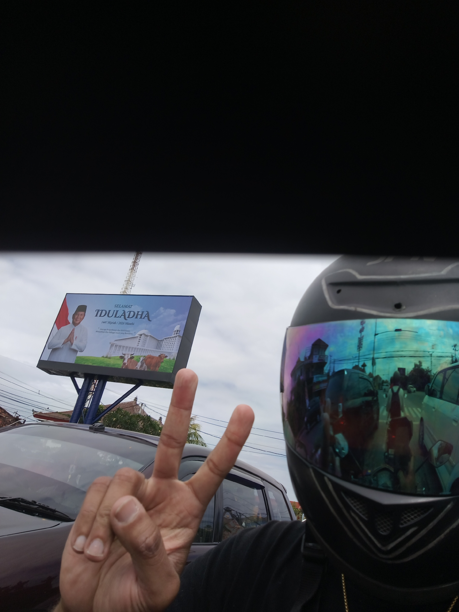 A selfie is captured from the perspective of a person wearing a black motorcycle helmet with a reflective visor, which shows a distorted view of the surrounding environment. The person is making a peace sign with their left hand, holding up two fingers. They are wearing a black t-shirt and a gold chain. In the background, a billboard displays "SELAMAT IDULADHA" along with an image of a man in traditional attire, a building resembling a mosque or government building, and two cows. The billboard is supported by a blue metal structure. To the left of the billboard, a cell tower is visible. In the foreground, the right side of a dark-colored car can be seen. The reflection in the helmet visor shows a street scene with other vehicles, buildings, and trees under a cloudy sky. The image appears to have been taken in an outdoor urban setting.