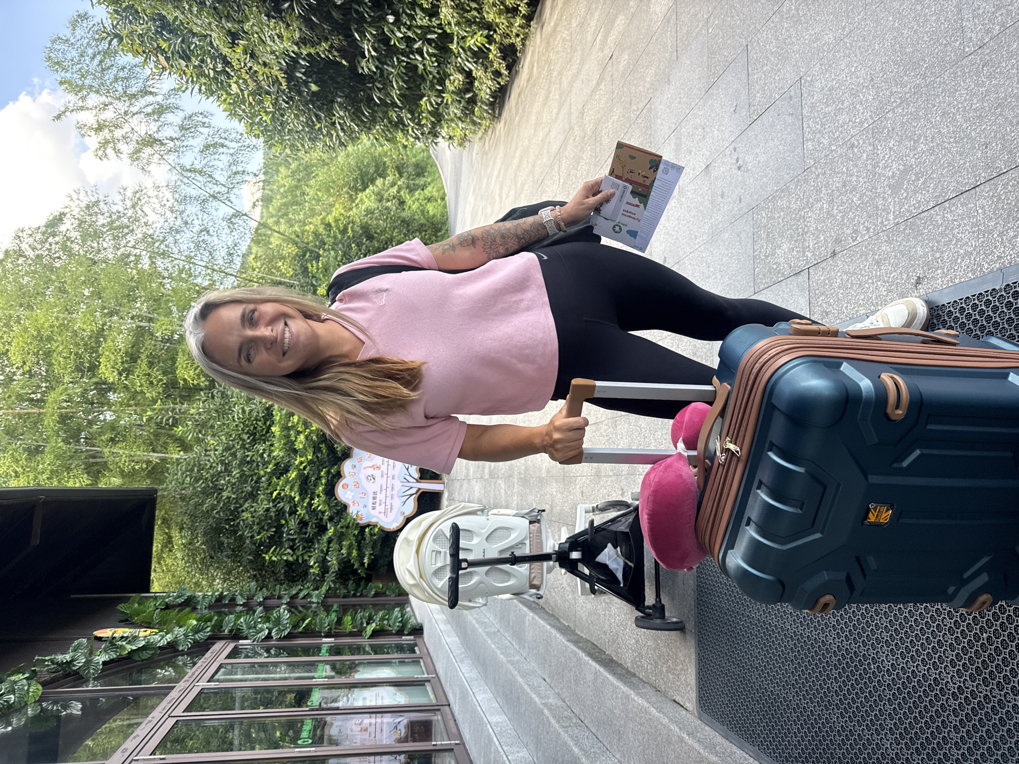A woman stands on a paved walkway in front of a building with large windows and a dark overhang. She is holding the handle of a dark teal suitcase and carrying a black backpack. In her right hand, she holds two pamphlets. She is wearing a pink v-neck t-shirt and black leggings. To her left, a white baby stroller is parked. Lush green trees and bamboo stalks are visible in the background, suggesting an outdoor, possibly park-like setting. The lighting indicates it is daytime. The building appears to be an entrance to a facility, possibly a tourist attraction or hotel, given the signage and landscaping.