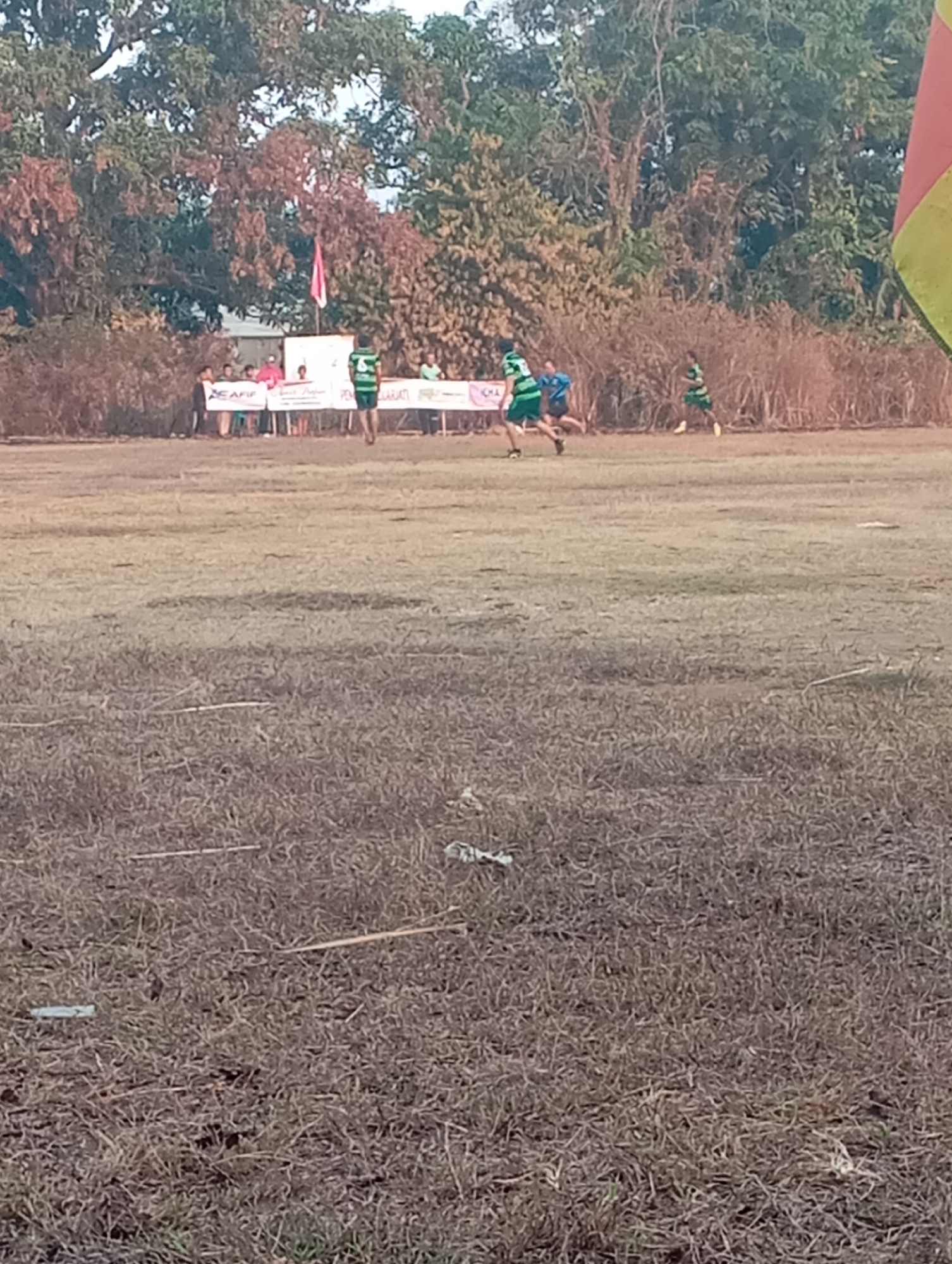 A wide shot shows a grassy field with several people in the background and some trees. The field is dry and sparse, with patches of brown grass. In the background, there is a group of people standing behind a banner, and several individuals are playing on the field. One person in the foreground is walking away from the camera. The sky is bright, suggesting it is daytime. The image is not clear enough to provide specific details about the people's clothing or facial features, or to discern the exact location within Kecamatan Haurgeulis, Indonesia.