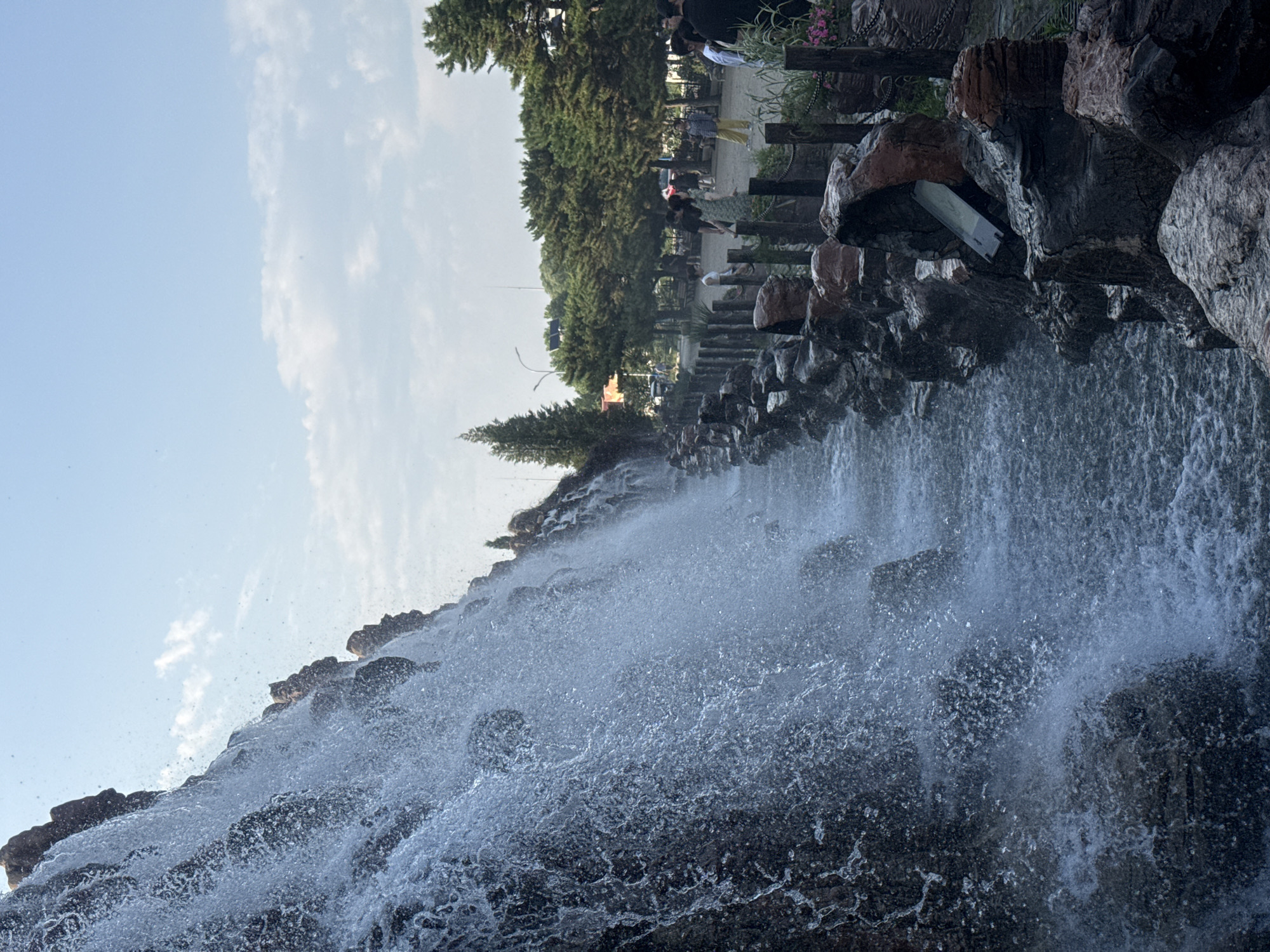 The image depicts a man-made waterfall cascading down a rocky incline. The water flows into a channel bordered by a rocky embankment. Beyond the embankment, a pathway is visible with several individuals walking and standing. Trees line the background, and a clear sky with scattered clouds is overhead. The scene appears to be an outdoor public space, possibly a park or a scenic area. The location is identified as Qinhuangdao, China.