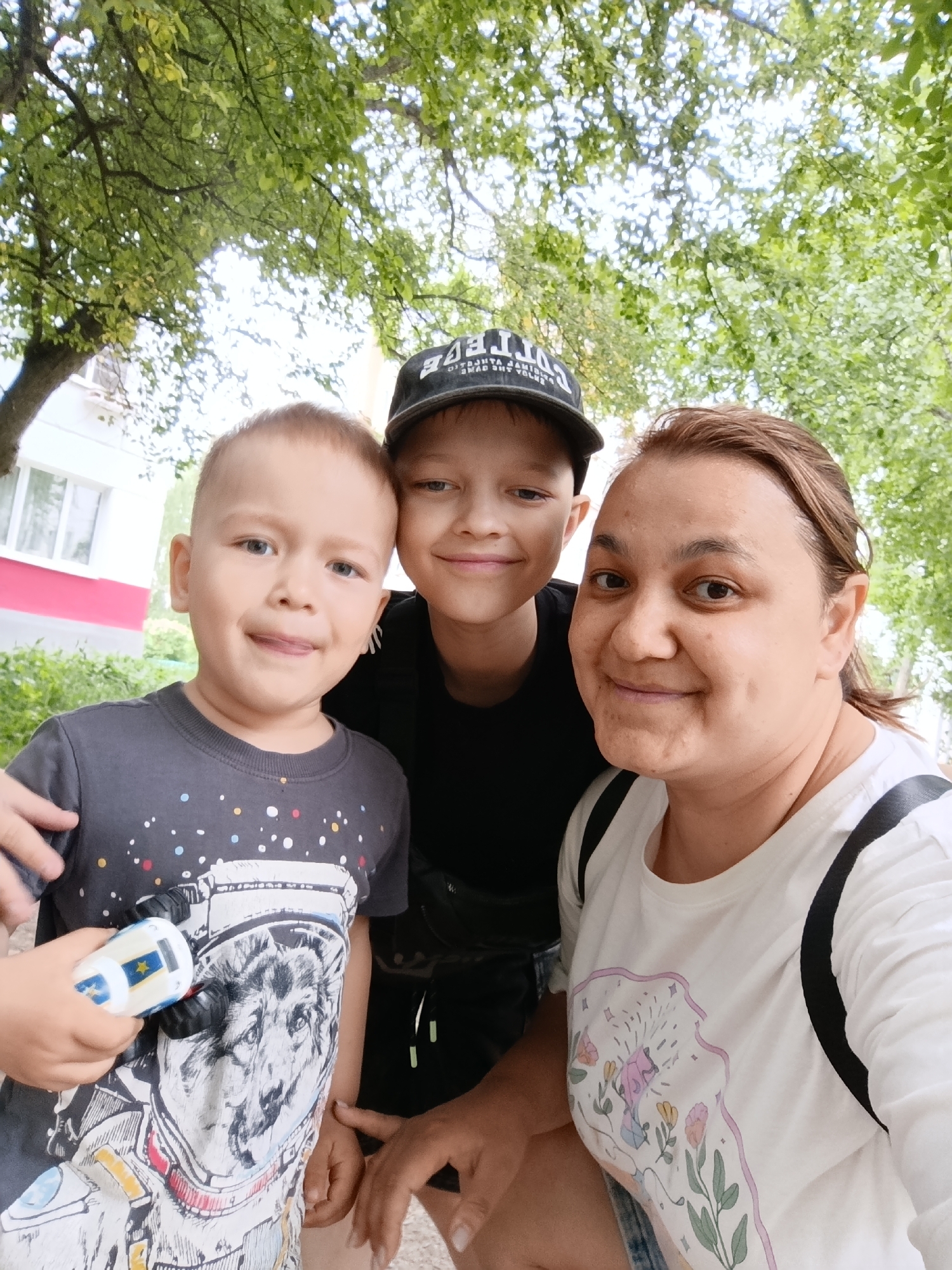 A woman is taking a selfie with two boys under a tree on a sunny day. The location is in the city of Chaikovsky, Russia. The younger boy is on the left, wearing a gray t-shirt with a wolf astronaut design and holding a toy car. The older boy is in the middle, wearing a black t-shirt and a black baseball cap. The woman is on the right, wearing a white t-shirt with a floral design and a backpack. Green trees and a building with a white window frame and red siding are visible in the background.