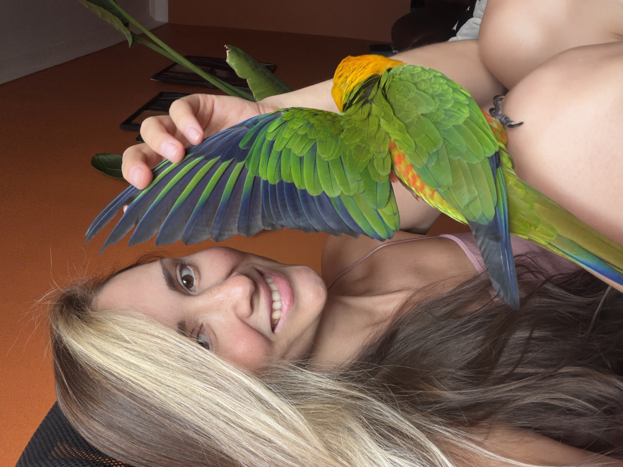 In this medium shot, an upside-down young woman with light brown and blonde hair lies on her back, her face turned towards the camera and smiling. She is wearing a pink tank top. Next to her, a bright green parrot with an orange-yellow head and blue-tipped wings is perched on her arm, its wing extended. The parrot appears to be nestled into her chest or shoulder. The background shows an orange-colored surface, possibly a floor or table, with some green leaves and a dark rectangular object, suggesting an indoor setting with a plant. The lighting suggests daytime. The overall mood is relaxed and affectionate, with the woman's smile and the close proximity of the parrot indicating a bond between them. The image appears to have been taken in Khimki, Russia. There is no visible text in the image.