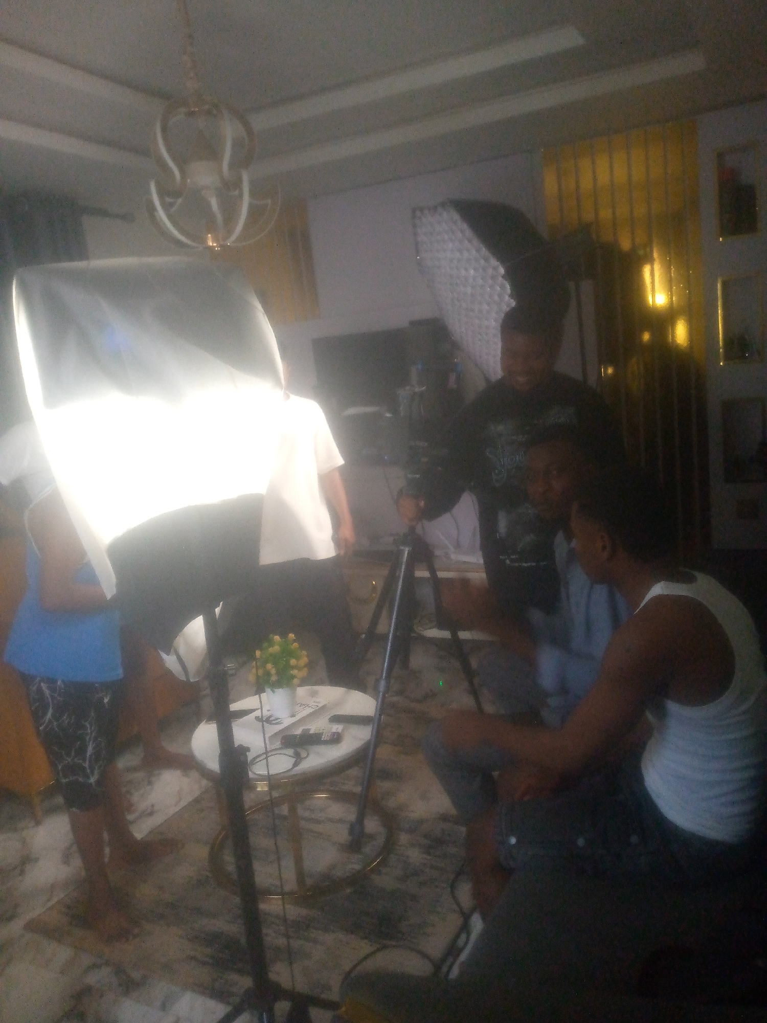 A group of people are inside a room with film equipment.
Several individuals are clustered around a tripod-mounted camera. One person is standing behind the camera, another seated in the foreground facing away from the camera, and another person in a white shirt visible behind the camera. A fourth person is standing to the left with their arms crossed. The foreground is dominated by a large softbox light. A small round table holds electronic devices and a decorative floral arrangement. In the background there is a decorative gold screen, a ceiling mounted chandelier, and a television. The patterned rug and other details suggest this is a residential indoor location.
The location, Ikorodu, Nigeria is provided as contextual information; there is no specific information within the image to independently confirm it.