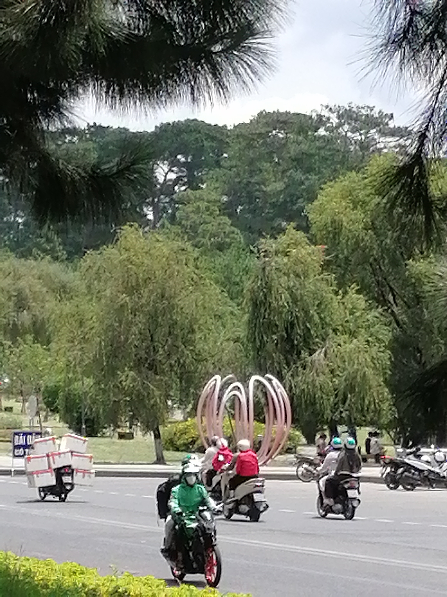 The image captures a street scene in Da Lat, Vietnam, characterized by lush greenery and a bright sky. In the foreground, a person wearing a green jacket and a mask is riding a black motorcycle with red accents on the wheels. The motorcycle is equipped with a black passenger seat and a black storage box behind the rider. To the left, another motorcycle is laden with a stack of white boxes, secured with straps. This motorcycle has a black seat and is being ridden by a person wearing a green helmet. Further in the background, several other motorcycles are visible, some carrying passengers. One motorcycle has two individuals wearing light-colored helmets and jackets, while another person on a motorcycle wears a red jacket and white helmet. A striking pink metallic sculpture, resembling an abstract flower or orb, stands prominently in the middle ground, adding an artistic element to the urban landscape. The background is filled with dense trees and foliage, suggesting a park or a tree-lined avenue common in Da Lat. A sign with Vietnamese text is partially visible on the left side of the road. The overall atmosphere appears to be a typical day in this Vietnamese city, with motorcycles serving as a primary mode of transportation for both individuals and deliveries.