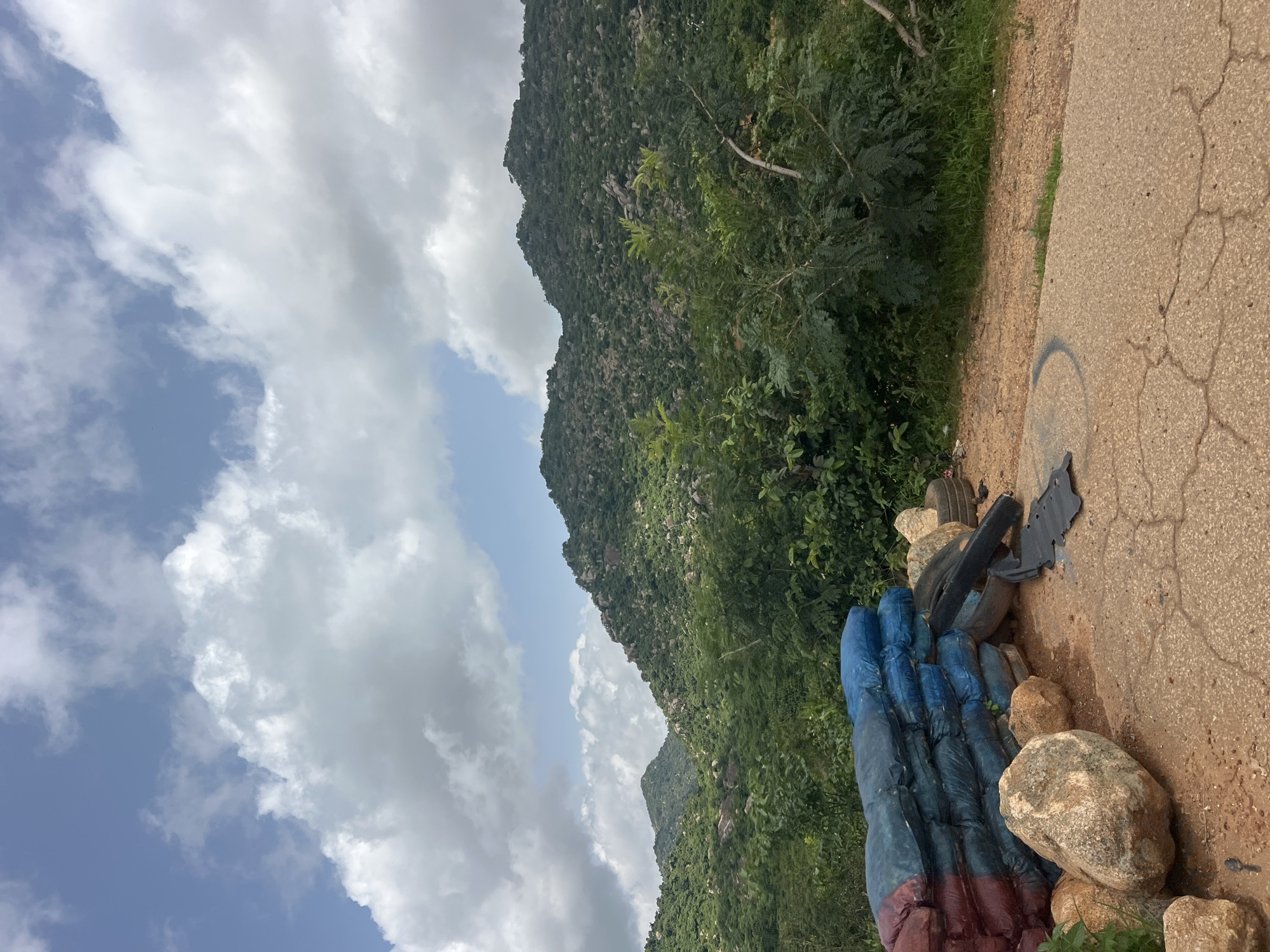 The image shows a roadside scene in Dorowa, Nigeria, featuring a pile of sandbags in blue and red, some large rocks, and discarded tires. A dark, possibly rubber mat-like object is on the ground near the tires. The foreground is a cracked asphalt surface with some gravel and dirt. Behind the sandbags and rocks, lush green vegetation and trees are visible, leading up to a series of rolling, rocky hills covered in dense greenery. The sky above is partly cloudy with patches of blue sky and white cumulus clouds. The overall impression is a natural landscape with man-made debris present.