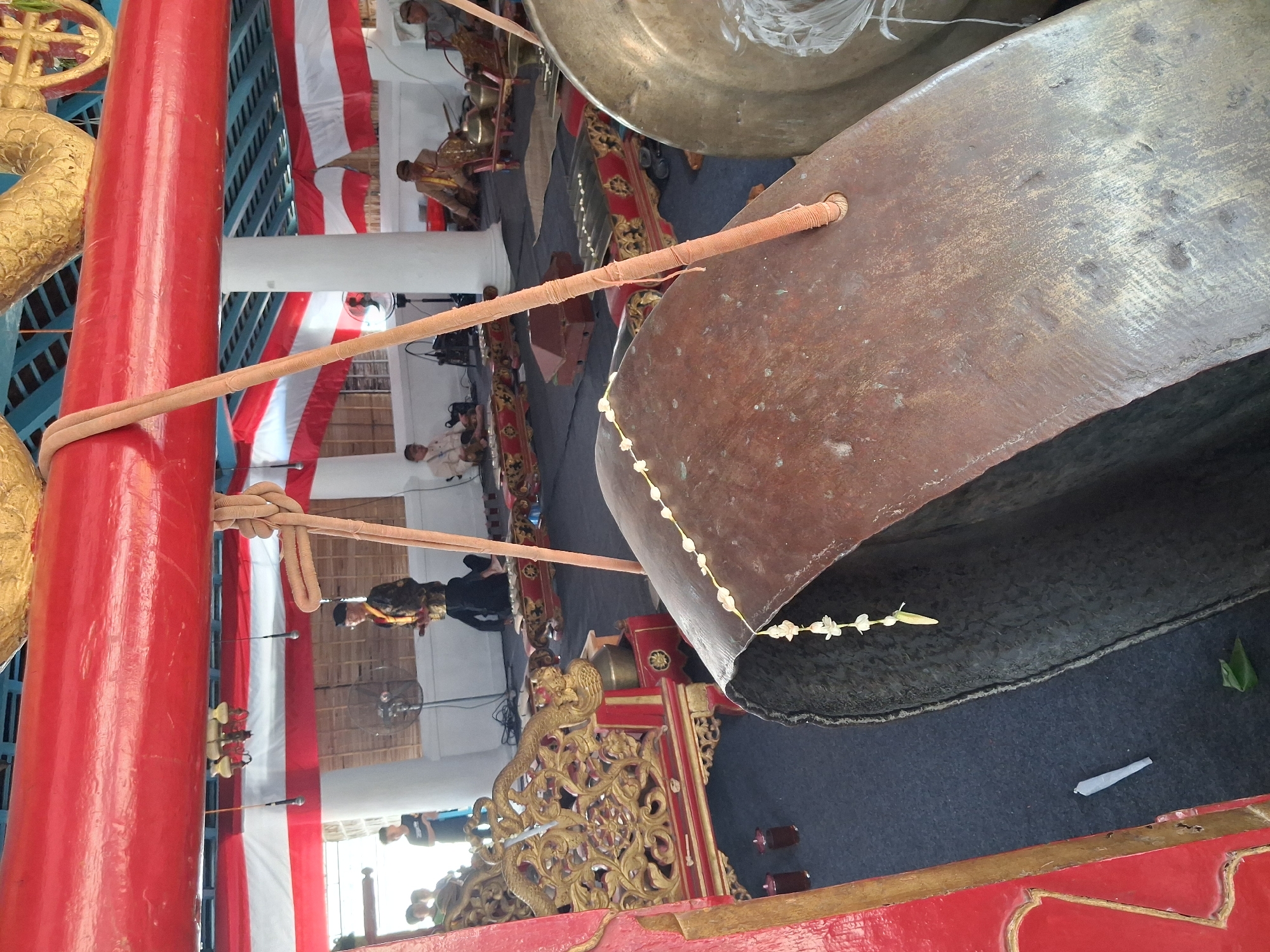 The image captures a scene within what appears to be a traditional Javanese performance space, likely in Surakarta, Indonesia, given the cultural context suggested by the Gamelan instruments. In the foreground, a large, weathered bronze gong is prominently featured, partially obscuring a red, ornate wooden structure that frames the left side of the image. A string of white flowers with a yellow center is draped across the gong.
In the mid-ground, several individuals are seated on the floor, arranged around Gamelan instruments. To the left, two individuals are visible, one in a black shirt and the other in a lighter colored shirt. A man in a traditional Javanese uniform, featuring a dark jacket with gold embellishments and a headdress, stands in the middle ground. He is holding a small object. Further back, three more individuals are seated, all appearing to be men, dressed in traditional attire, and positioned near Gamelan instruments. One of these men is playing an instrument.
The background reveals a wall covered with woven bamboo blinds, and above it, red and white striped banners are hung. The ceiling structure is visible, with a large, red-painted beam spanning across the upper portion of the frame. The floor is covered with a dark carpet. In the extreme foreground, a single white cigarette is placed on the carpet, next to a green leaf.