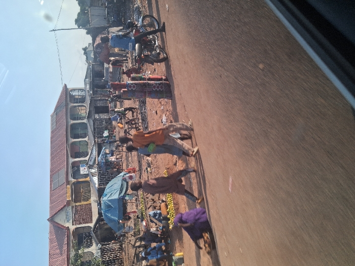 This image captures a vibrant, open-air market scene along a dirt road in Mutum Biyu, Nigeria, under clear daylight. The perspective is from inside a vehicle, indicated by a dark frame in the lower right corner.
In the foreground, a reddish-brown dirt road runs horizontally. On the left side of the road, a person in a purple garment is seated on the ground, attending to a shallow, dark pan containing items. Three individuals walk across the road from left to right: one young person in a brown garment, followed by another in light blue, and a third in an orange shirt and light trousers. To the right, a person in a blue shirt is seated on a red motorcycle, which is parked facing right on the roadside. Nearby, stacks of colorful, patterned cylindrical items are vertically arranged on the ground.
The midground is dominated by a bustling market area situated to the right of the road. This area features multiple makeshift stalls with corrugated metal roofs and at least one prominent blue tarpaulin tent providing shade. Various individuals are visible within the market, sitting, standing, and interacting. Piles of goods, specifically numerous yellow citrus fruits, are displayed on the ground near vendors. The market ground is a reddish-brown dirt surface, scattered with debris.
In the background, a two-story building with a red corrugated metal roof and a light-colored facade stands out. This building features several arched windows or balconies, and solar panels are installed on its roof. A partially legible sign on its facade appears to read "SELIMAZ" or similar. To the right of this building, other less distinct structures are visible. A utility pole with overhead wires is present on the far right. The sky above is clear and light blue.