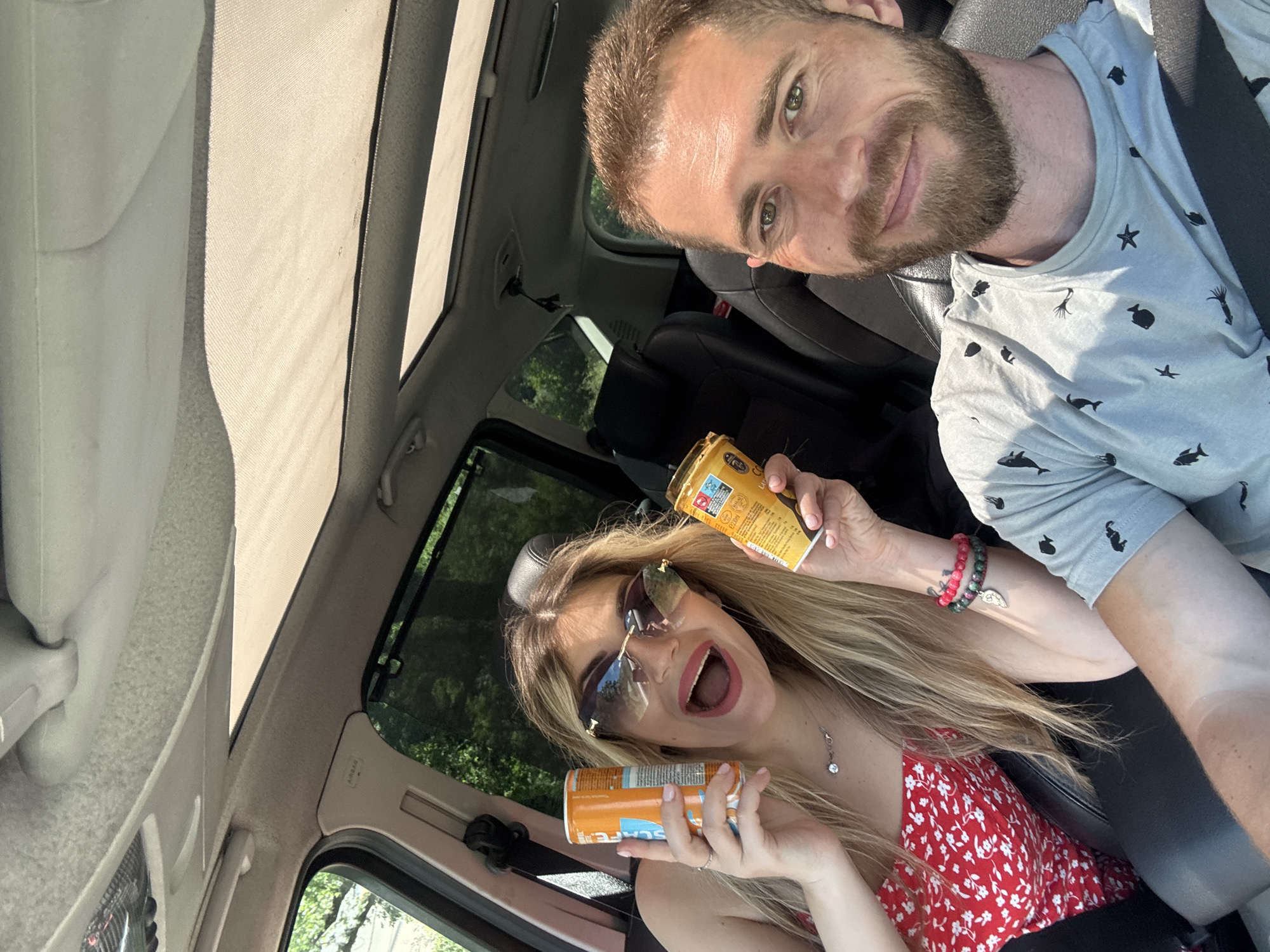 A bright, daytime selfie captures a man and a woman inside a car, likely on a journey through Kaluđerac, Montenegro. The image is taken from a high angle, looking down into the vehicle, and is rotated, with the man's head at the top and the woman's at the bottom.
The man, on the right, has a beard and short hair, and wears a light grey t-shirt adorned with black marine animal patterns including fish, starfish, and squids. He is smiling faintly, looking directly at the camera, with a seatbelt fastened across his chest. His forehead shows some sun glint.
The woman, on the left, has long blonde hair and is wearing a red and white floral top. She has dark sunglasses perched on her head and a wide, open-mouthed expression of excitement or surprise. She wears a silver necklace with a small pendant and beaded bracelets on her left wrist, one of which features a small wing charm. She holds two canned beverages. The can in her right hand is golden-brown with "ICED COFFEE" and "LATTE" clearly visible. The can in her left hand is orange and shows the brand name "E-SCAPE."
The car's interior details include a light-colored headliner, a visible sunroof allowing bright sunlight to flood the cabin, and black leather seats. Green foliage from trees is seen through the car windows, suggesting a natural or scenic outdoor setting. The strong, direct sunlight indicates a clear and bright day, contributing to the cheerful and energetic mood of the subjects.