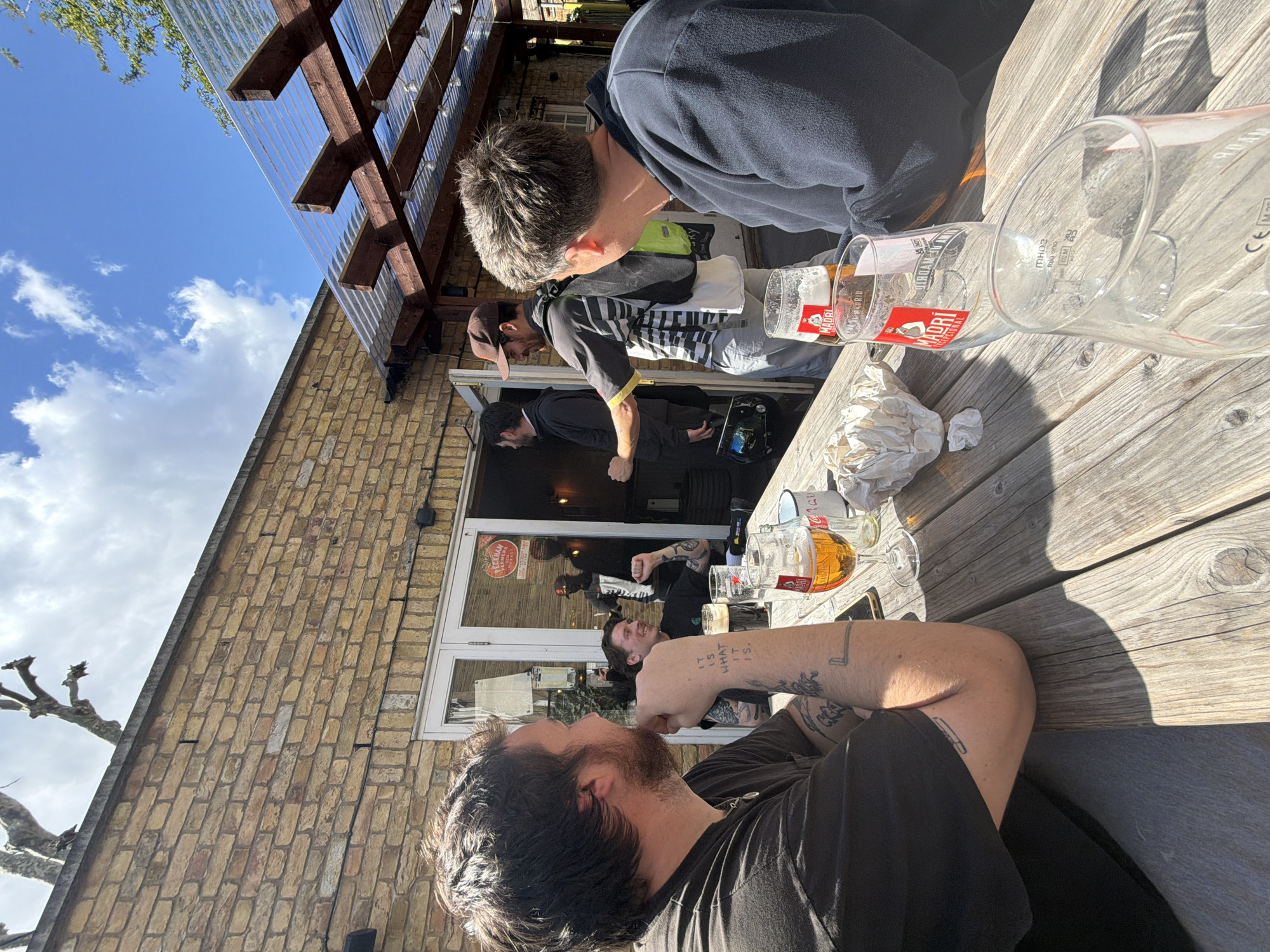 The image depicts a group of people at an outdoor seating area of a building in London, United Kingdom. The building appears to be a pub or restaurant, constructed of light brown bricks. There is a covered roof section overhead, with a view of the sky and clouds above. Several individuals are seated at a wooden table, where a few beer glasses, some bearing the "MADRI" logo, are visible. One of the people at the table has visible tattoos on their forearm. Through the doorway of the pub, other people are seen inside, with some standing. The atmosphere suggests a casual social gathering.