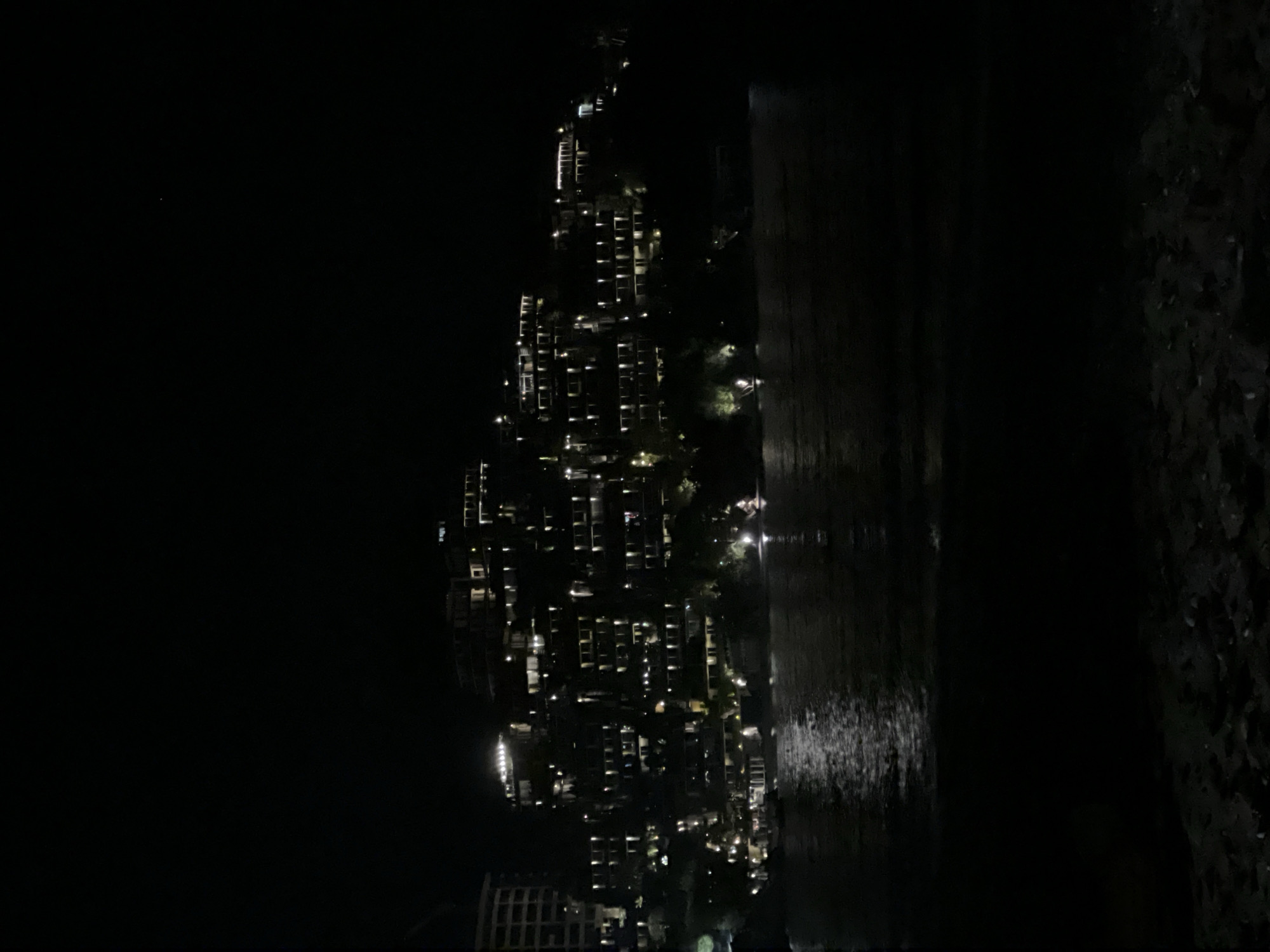The image shows a nighttime view of a high-rise building in Budva, Montenegro, as seen from the beach. The building is illuminated with numerous lights, indicating residential or commercial occupancy. The water reflects some of the building's lights. To the right, a dark, textured area indicates a rocky or vegetated coastline. The image is mostly dark, with the building's lights providing the primary source of illumination.