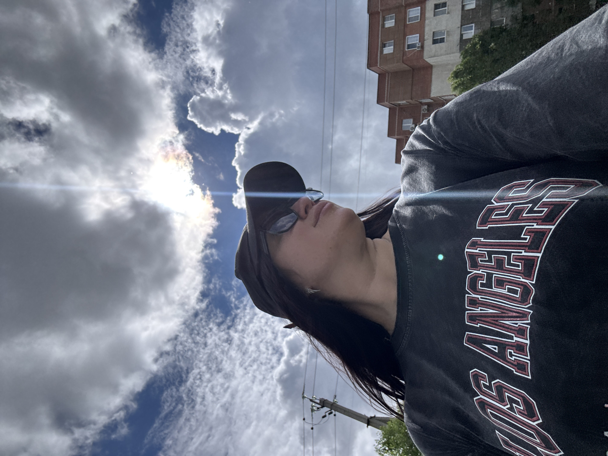 A low-angle, rotated photo depicts a person, likely a young woman, from the chest up, looking upwards towards a bright, partly cloudy sky. She is seen in profile, wearing a black baseball cap and sunglasses, with long dark hair visible beneath the cap. Her dark grey or black t-shirt features large, outlined text in white and red, partially spelling out "LOS ANGELES".
The sky is a mix of deep blue patches and large, white and grey clouds. The sun is intensely bright, visible behind some clouds, creating a prominent lens flare across the image. In the background, a multi-story building with numerous windows, possibly an apartment or office complex, stands against the sky. Utility poles and overhead wires are also discernible. The photo's orientation is rotated 90 degrees clockwise, placing the subject horizontally. The setting appears to be outdoors during the daytime, likely in an urban or suburban environment. The person's expression is obscured by her sunglasses and the angle of the shot, suggesting she might be shielding her eyes from the sun while observing the sky.