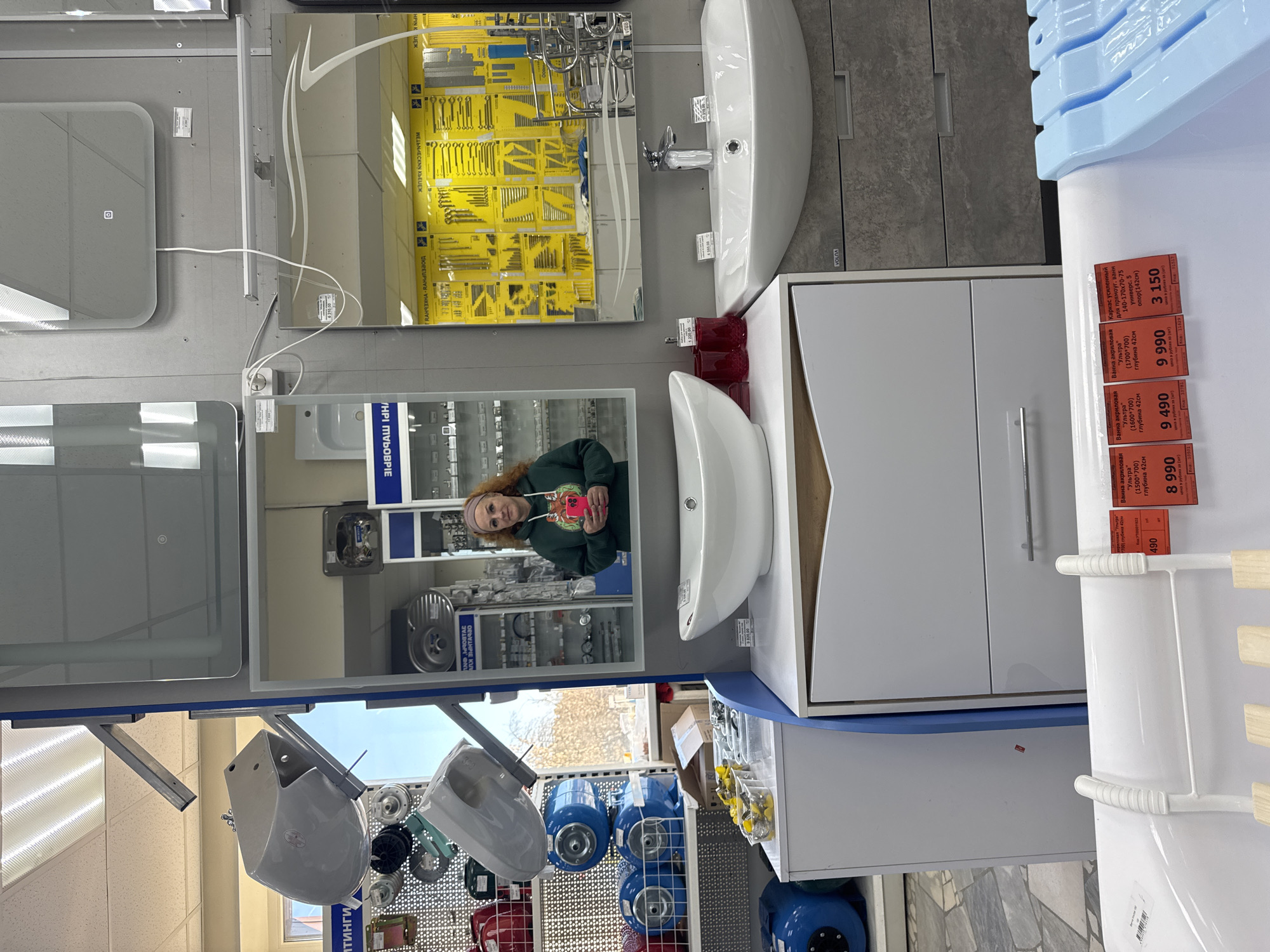 The image appears to be taken inside a plumbing supply store in the city of Chaikovsky, Russia. A woman with red hair and a gray headband is visible in the reflection of a mirror, holding a pink phone. The scene includes bathroom fixtures such as sinks, vanities, and mirrors displayed on shelves. The items are priced in rubles and labeled in Russian, referencing bathroom fixtures, acrylic bathtubs, and reinforced frames. The store also appears to stock plumbing parts and tools.