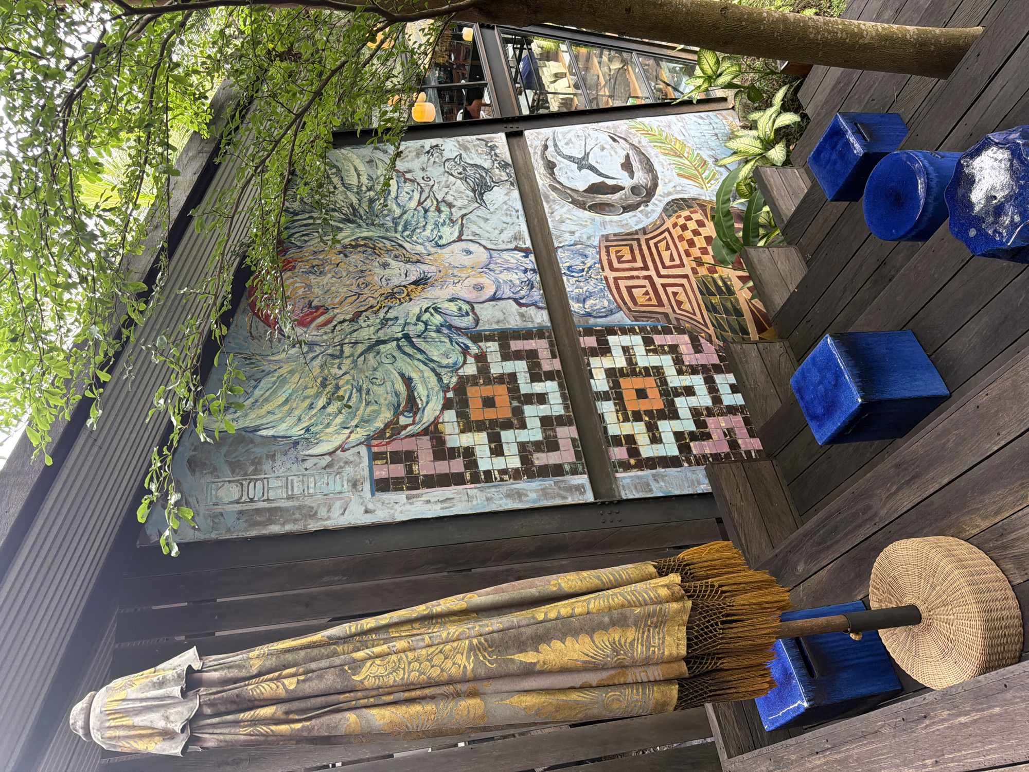 The image depicts an outdoor seating area with a striking mural and an umbrella. The mural, a large, colorful artwork, features a winged figure, a bird, and celestial imagery. In front of the mural, a closed umbrella with an ornate gold pattern and fringe stands on a wicker base. Several bright blue ceramic stools of various shapes are arranged on a wooden deck, which appears to be part of a multi-level outdoor space. Green foliage from a nearby tree drapes into the scene, and the background reveals glimpses of an indoor space with lighting fixtures and seating through large windows. This setting is identified as being within the city of Sukawati, Indonesia.