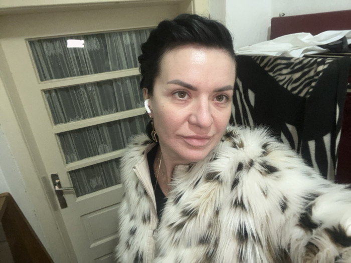 This image depicts an adult female in an indoor setting within Smederevska Palanka Municipality, Serbia. The subject, appearing to be in her 40s or 50s, has dark hair, styled short or swept back. She is looking directly at the camera with a neutral expression, taking a selfie. She wears a multi-toned faux fur coat, primarily white with black and brown markings, over a light-colored jacket and a black garment. A white wireless earbud is visible in her right ear, along with a gold-colored hoop earring and a thin gold-colored necklace.
The background to the left features a light-colored wooden door with multiple rectangular glass panels, each covered by white sheer curtains or vertical blinds. A silver-toned door handle is visible. To the right, a piece of furniture, possibly a chair or headboard, has a visible dark red/maroon section at its top edge. A black and white zebra-print blanket or cover is draped over the lower part of this furniture. The overall lighting is consistent with an indoor environment.