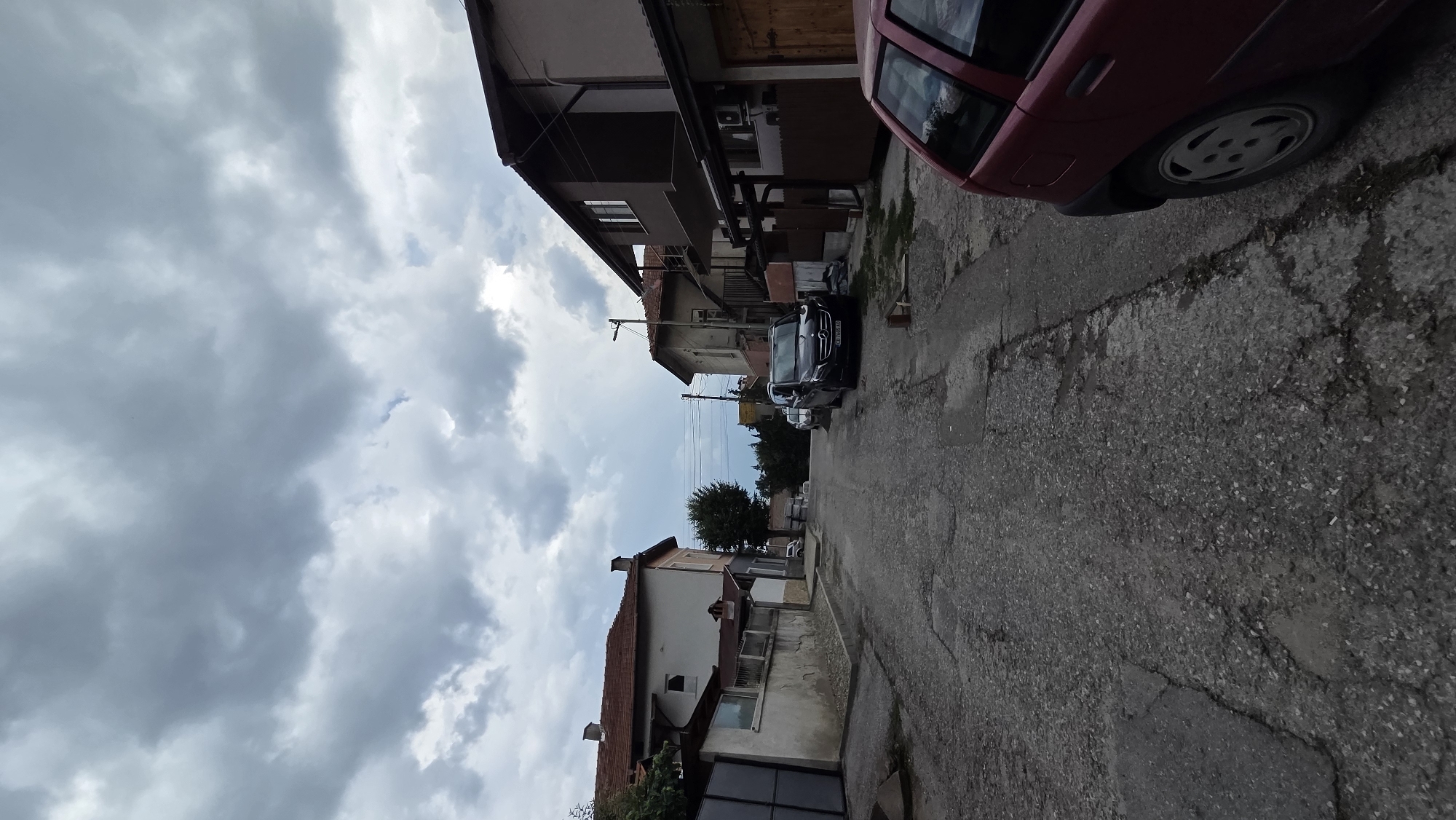 The image depicts a view looking down a narrow, paved street in the city of Bania, Bulgaria, under a cloudy sky. The street is in poor condition, with visible cracks and potholes. On the left side of the street, a white building with a red-tiled roof and a grey fence is visible, with a large metal gate. Further down the street, a lone tree stands between two buildings.
On the right side of the street, a multi-story building with a dark gray facade and a red-tiled roof is prominent. A black Mercedes-Benz van is parked on the right side of the street. In the foreground on the right, the rear of a red Fiat Punto is visible. The environment appears to be a residential area, with evidence of aging infrastructure. No people are clearly visible in the scene. The overall atmosphere is subdued due to the overcast weather.