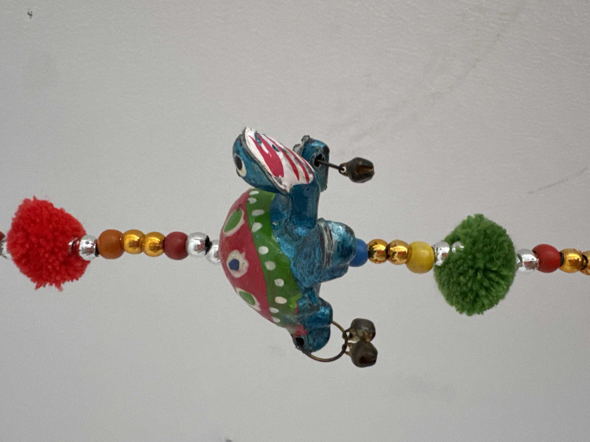 A close-up, eye-level, vertical shot showcases a colorful, handcrafted wind chime suspended against a pale, textured wall, likely within a home in Valencia, Spain. The primary subject is a whimsical, blue turtle figurine, its shell adorned with a vibrant red, green, and white polka-dot pattern. Red and white stripes decorate its face, and its eyes are a striking blue. Dangling from the turtle's sides are two small, dark bells.
The turtle is part of a decorative strand composed of various beads. Above the turtle, a fluffy red pom-pom is attached, followed by a sequence of golden, brown, and silver beads. Below the turtle, a bright green pom-pom is interspersed with more golden and silver beads, with a single blue bead just above it. Further down the strand, a red bead and more golden beads are visible.
The lighting is soft and even, suggesting an indoor setting. The background is a plain, off-white wall with a subtle texture, devoid of any distinguishing features or external elements that would indicate a specific outdoor location in Valencia. The focus is entirely on the decorative object, highlighting its intricate details and playful design.