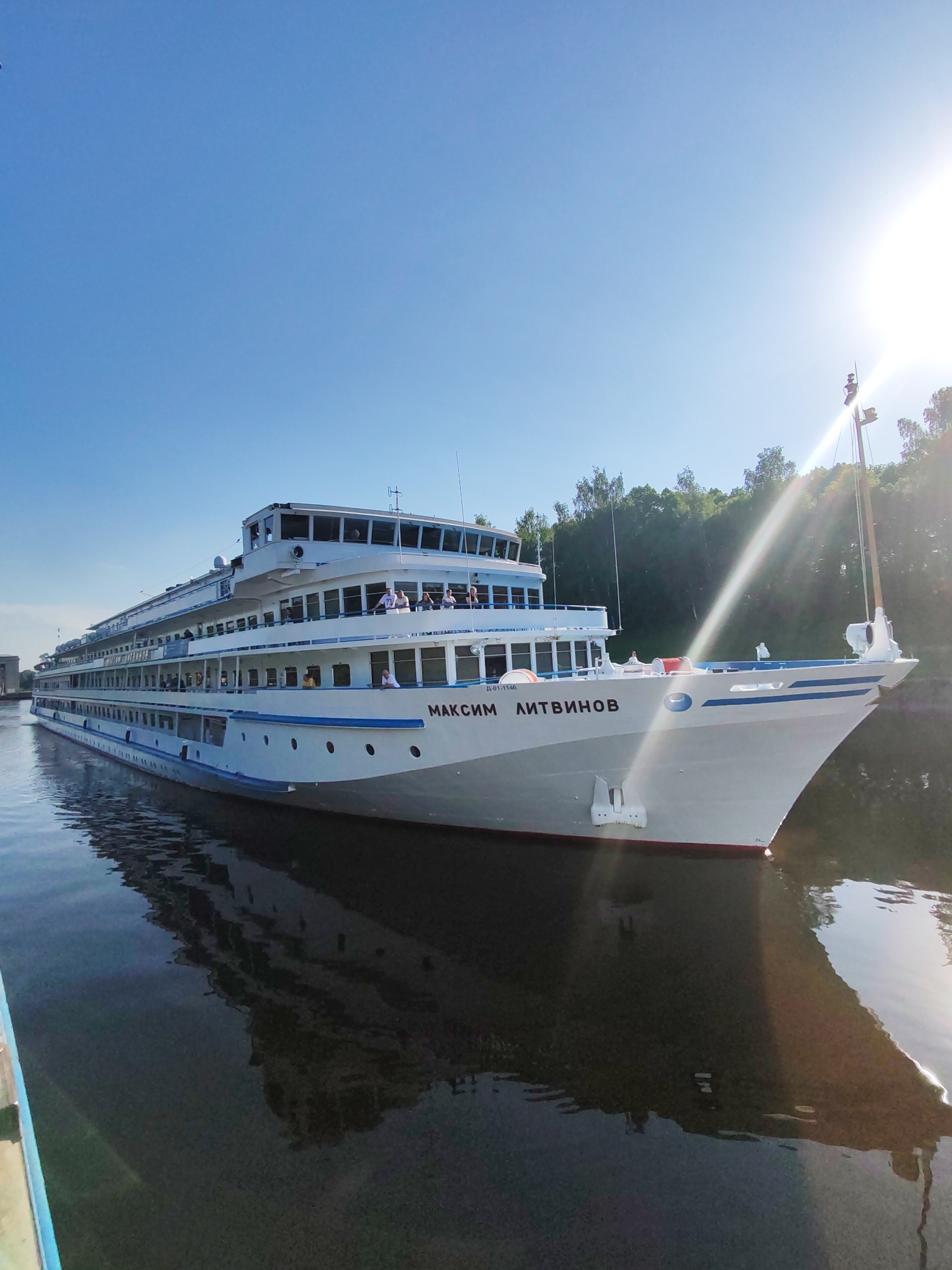 The image depicts the river cruise ship "Maxim Litvinov" sailing on a waterway in Iksha, Russia. The ship is white with blue trim and lettering and has multiple decks. People can be seen on the decks. The water reflects the ship and the clear blue sky. Green trees line the shore in the background. The sun is visible and casts a glare on the scene.