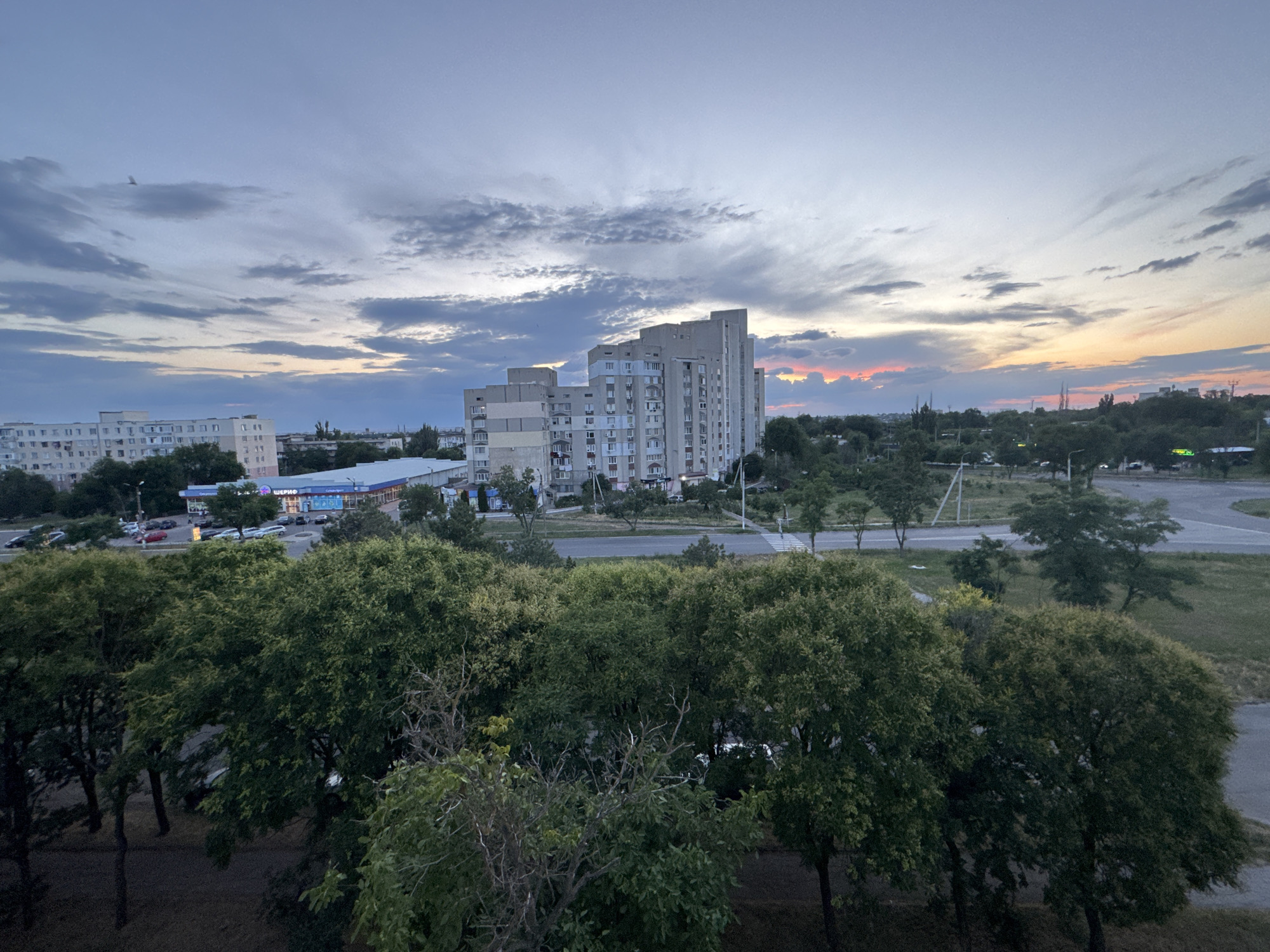 The image depicts an urban landscape at dusk, likely Tiraspol, Moldova, as indicated by the presence of Soviet-era architecture. The sky is a canvas of soft blues and greys, transitioning to warm hues of orange and pink at the horizon, suggesting the setting sun. A flock of birds is visible in the upper left portion of the sky. In the middle ground, a row of apartment buildings stands, characterized by their uniform concrete facades and numerous windows. These buildings are a common feature in post-Soviet urban planning. A street runs horizontally across the mid-section of the image, with a few cars parked and some in motion, indicating ongoing activity. A pedestrian crossing is visible on the right side of the street. In the foreground, a dense canopy of green trees dominates the scene, their leaves illuminated by the fading light. The overall impression is that of a quiet evening in a residential area, with hints of urban life continuing under the twilight sky.