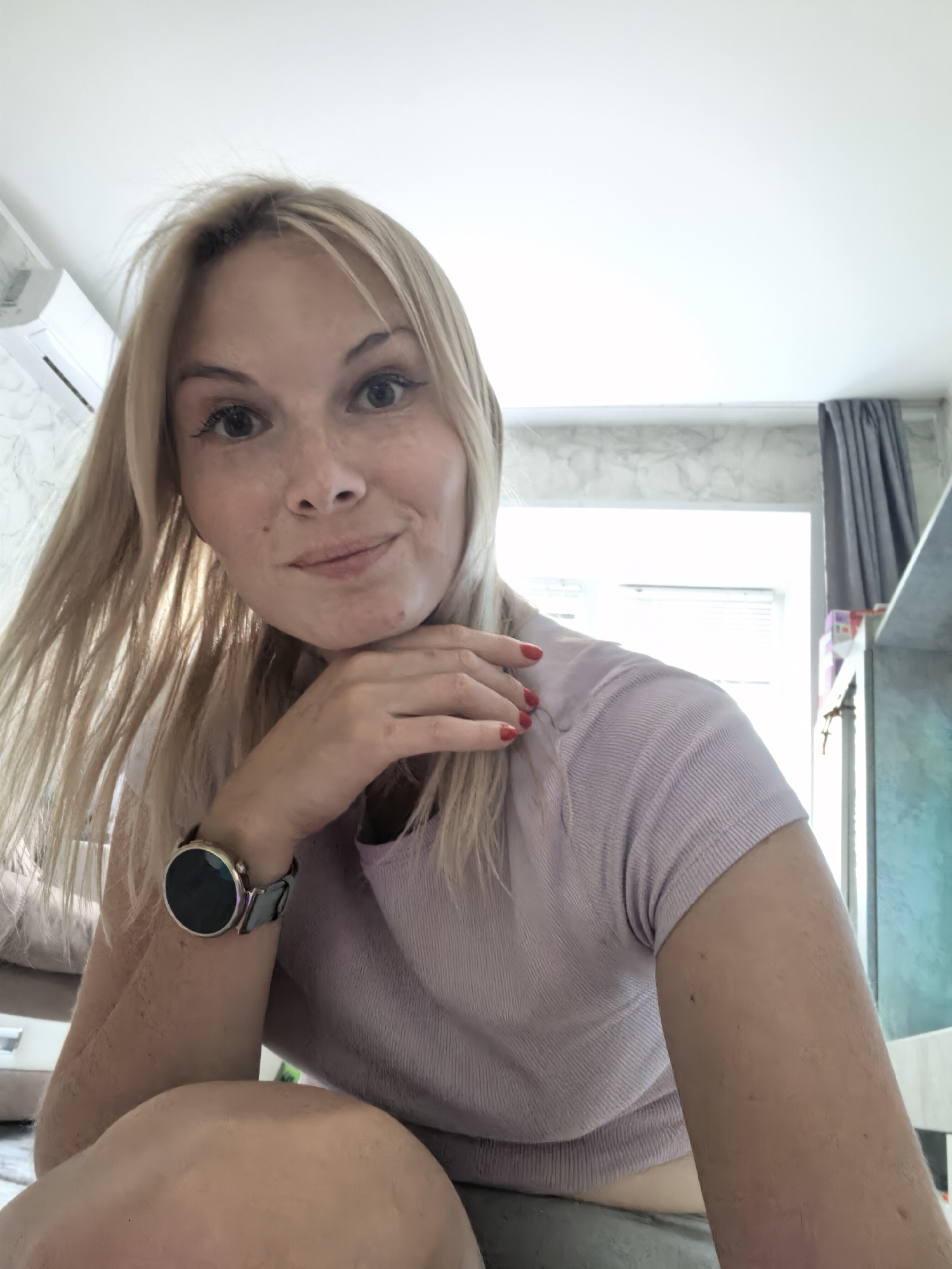 A fair-skinned woman with blonde hair and freckles is in a selfie, holding her chin with her right hand. She is wearing a light purple, ribbed t-shirt and a watch with a black band on her left wrist. Her fingernails are painted red. The background appears to be an indoor setting with white walls, a window with gray curtains, and what looks like an air conditioner unit. The lighting is bright, suggesting daytime. The location is not identifiable as Sterlitamak, Russia.