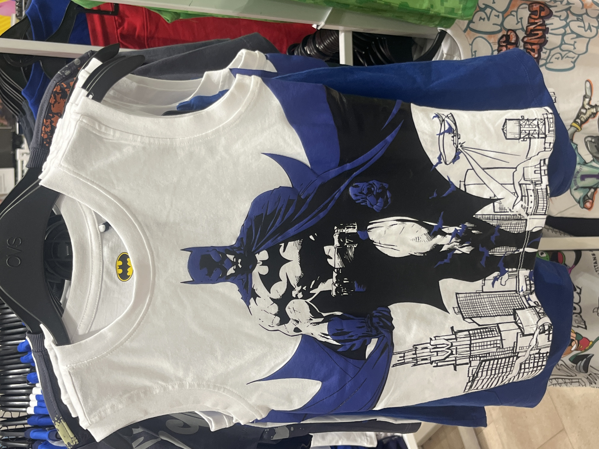 A white sleeveless t-shirt with a graphic of Batman is displayed on a clothing rack. The t-shirt features a large illustration of Batman in a dynamic pose, with a cityscape and a blimp in the background. The Batman logo is visible on the upper chest area of the shirt. The t-shirt is hung among other clothing items on black hangers. The scene is indoors, likely in a retail store. There is no indication of Vallo della Lucania, Italy in the image.