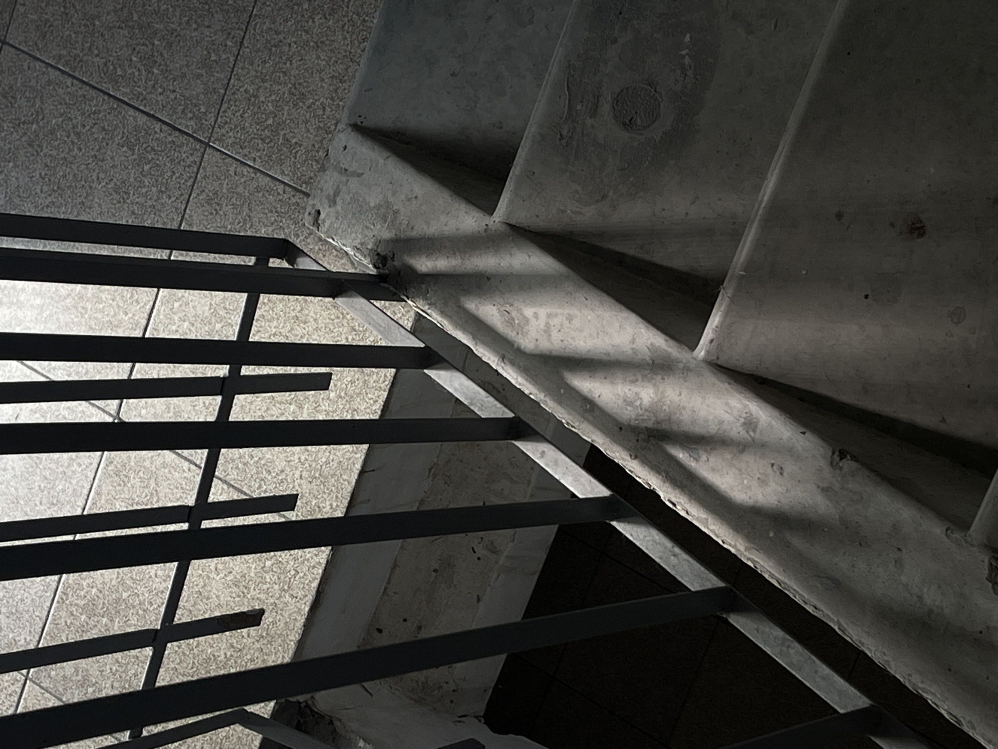 The image shows a concrete staircase with a metal railing. The stairs are outdoors, likely in a public space, as suggested by the tiled floor and the overall utilitarian design. Sunlight is casting shadows from the railing onto the stairs and the floor. The scene is devoid of people. The condition of the concrete suggests some wear and tear, with minor chips and imperfections visible. The overall impression is one of an ordinary, possibly aged, urban setting. The image does not contain any specific landmarks or clues that would place it in Rostov-on-Don, Russia, or any other particular city.