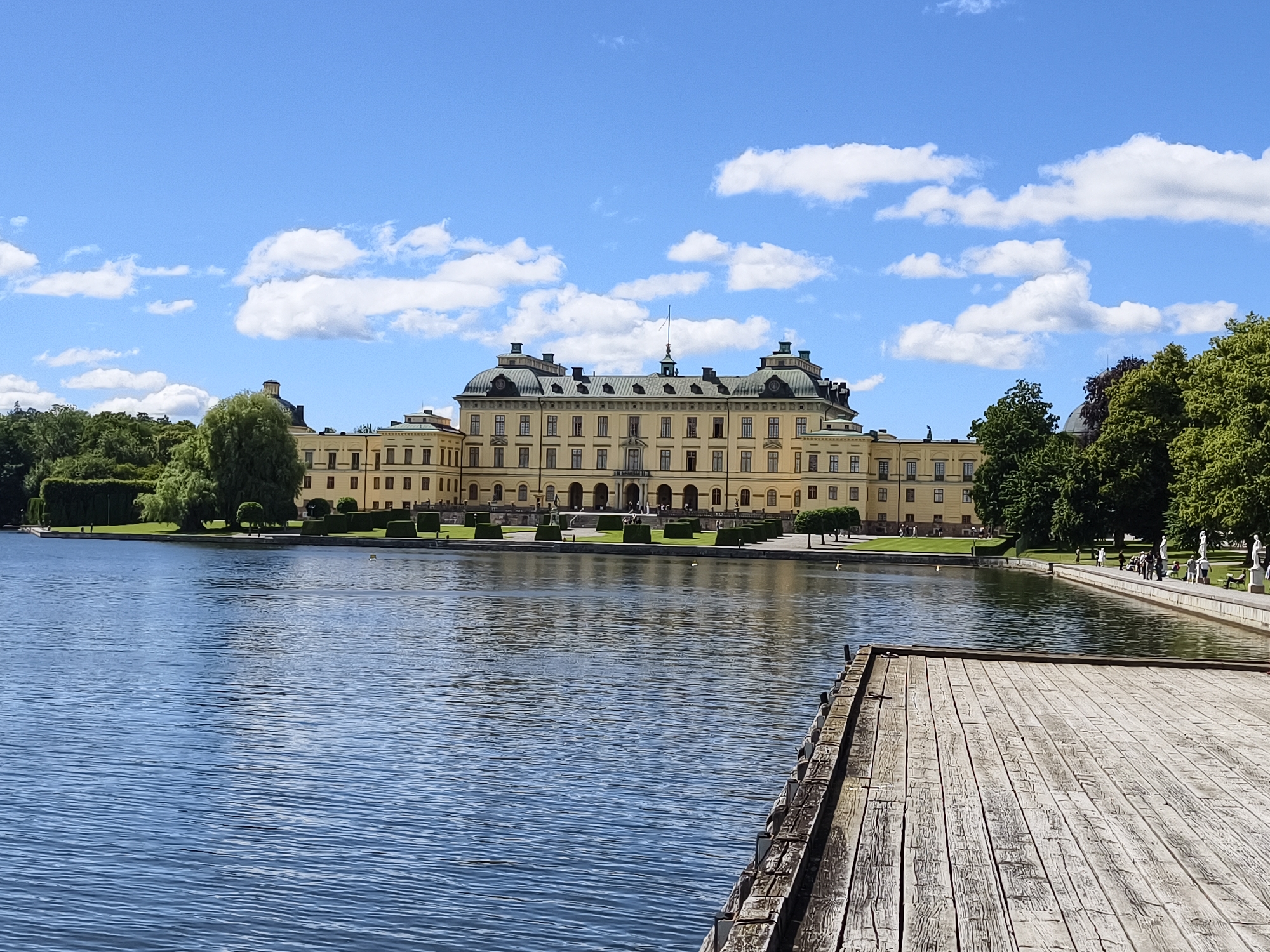 The image shows a long, yellow Baroque palace with a green mansard roof, situated behind a body of water. The palace has a symmetrical facade with numerous windows and several prominent domed structures. The water in the foreground is calm, reflecting the blue sky and scattered white clouds. A wooden dock extends into the water from the lower right corner of the frame. The grounds surrounding the palace appear to be well-maintained with manicured lawns, hedges, and trees. Several people are visible walking along a paved area near the palace and water's edge. The scene is bathed in bright daylight, suggesting it is daytime. This is Drottningholm Palace, located in Ekerö kommun, Sweden.