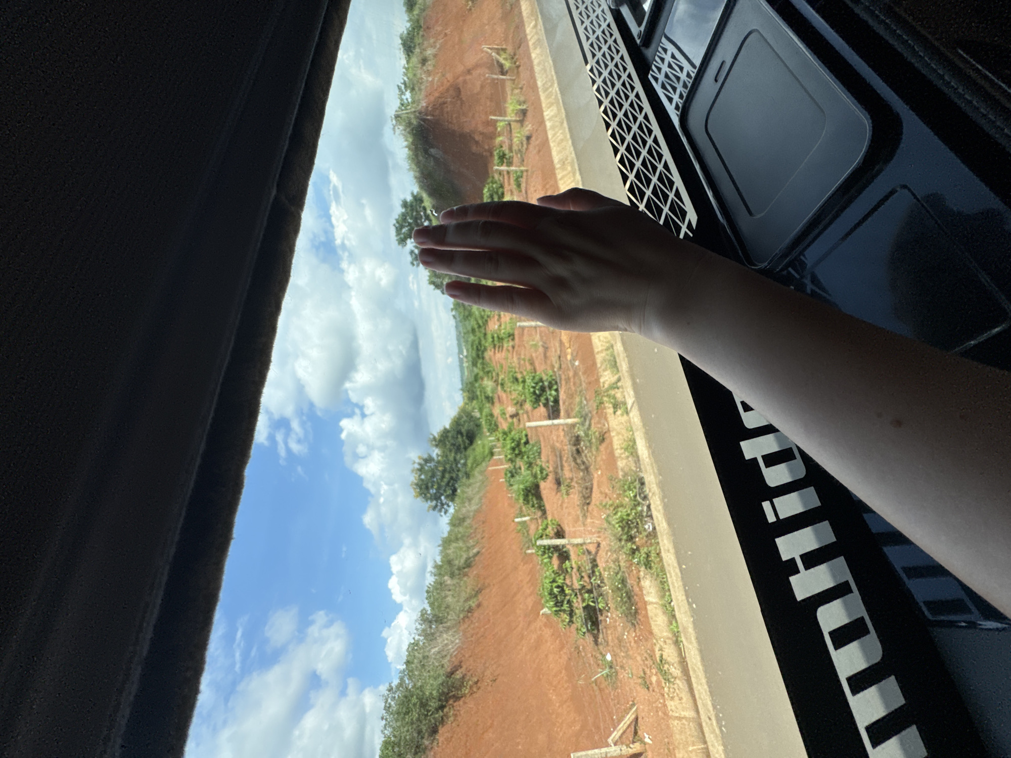 The image shows a person's left hand, palm facing forward, held up to a window. The hand is in the foreground, obscuring part of the view. Through the window, a landscape is visible, featuring a red, hilly terrain with scattered green vegetation and small plants planted in rows. The sky is blue with scattered white clouds. Below the window, a black surface with a white logo that appears to read "toHide" is visible. There is also a textured black grid pattern and a dark rectangular object on this surface. The scene suggests the person is inside a vehicle, possibly a truck, looking out at a rural area. The location is identified as Pleiku, Vietnam.
