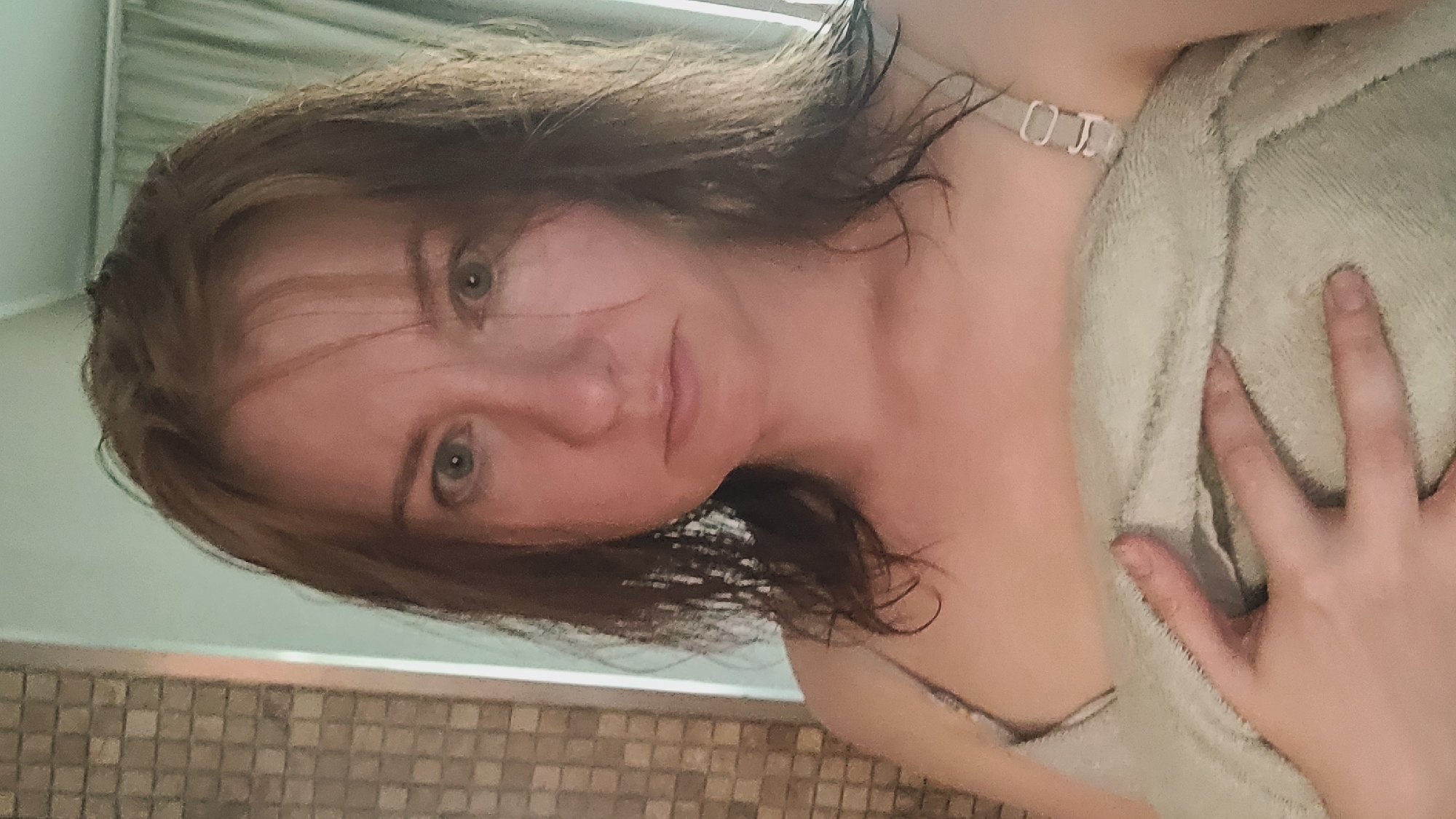 This media file is a close-up, portrait-style image of a person. The person appears to be a young adult with light skin and brown hair that is wet and somewhat disheveled, framing their face. Their eyes are open and looking directly at the camera, and they have a neutral to slightly soft expression. They are wrapped in a light-colored towel, which covers their body from the chest up. A hand is visible resting on the towel, with fingers slightly splayed. A delicate, silver-colored strap or necklace is visible around the neck. The background is blurred, but suggests an indoor setting with tiled walls or a textured surface below and a lighter, possibly curtained or paneled surface above. The lighting appears soft, possibly from an overhead source, creating some shadows on the face. The overall impression is intimate and personal, possibly taken in a bathroom or after a shower. No text is visible in the image.