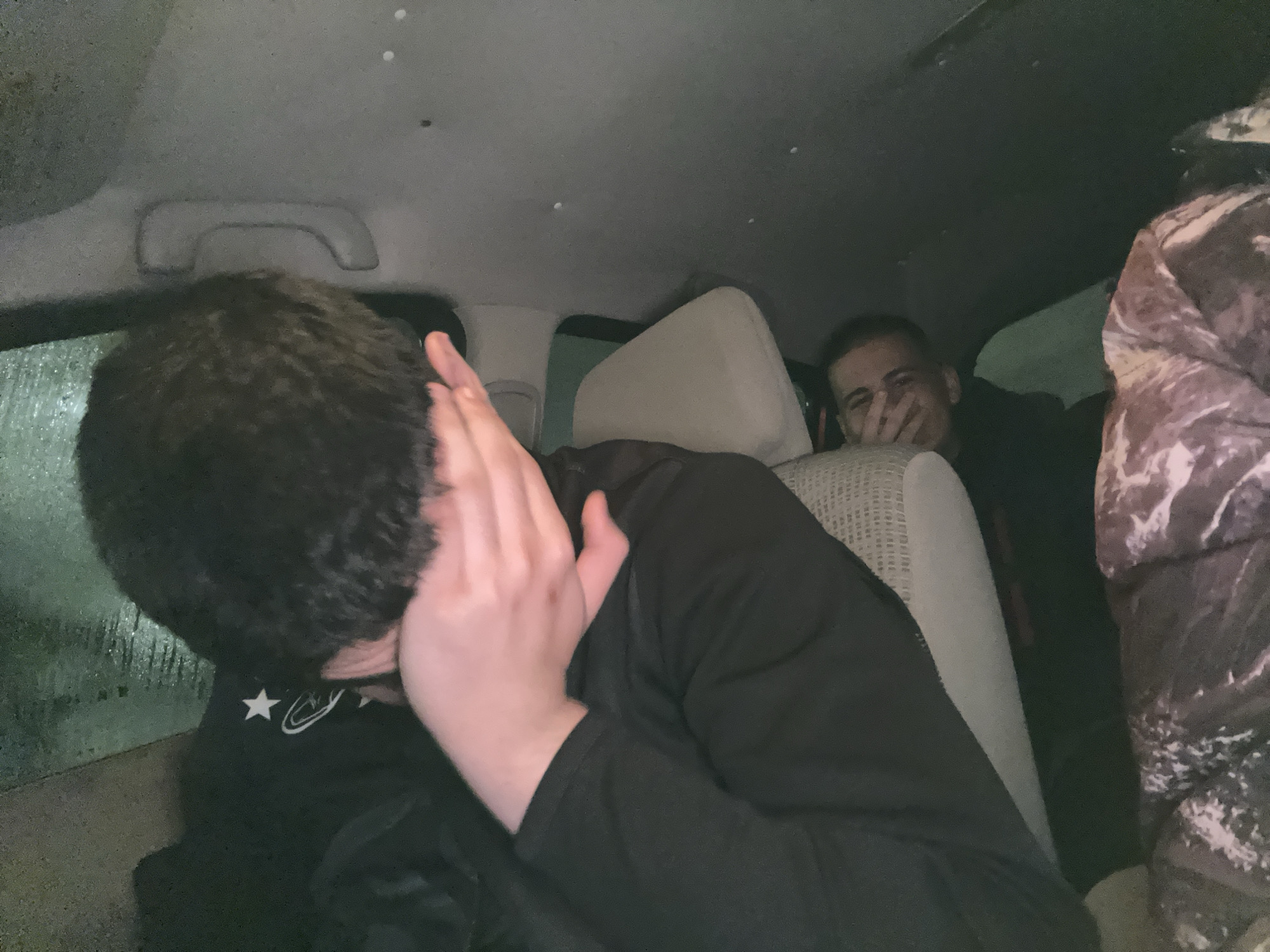 The image depicts the interior of a vehicle at night, in the city of Unknown, Unknown. Three individuals are present. In the left foreground, a person with dark, curly hair is seated, obscuring their face with both hands, palms inward. They wear a black long-sleeved garment featuring a white star and a white abstract circular design on the collar area. In the background, a second individual, a male with short hair and light skin, is partially visible behind a beige car seat, smiling or laughing with his right hand covering part of his mouth. To the far right, a third person's right shoulder, clad in a brown, white, and dark-patterned puffy jacket or blanket, is visible. The vehicle's interior includes a grey headliner with a visible grab handle. The window on the left displays numerous water droplets against a dark, greenish background, consistent with rain or condensation during nighttime or low-light conditions.