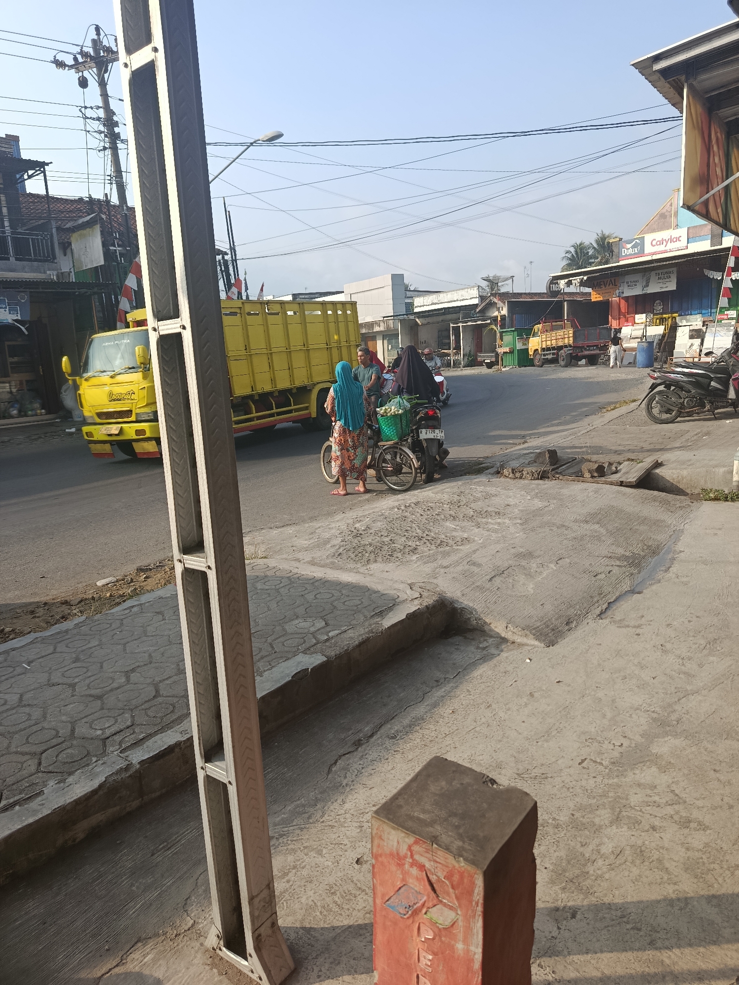 This image captures a street scene in Kalijeruk, Indonesia, on a sunny day. A yellow truck is parked on the right side of the road, and a woman wearing a blue hijab and a patterned dress is standing next to it with a bicycle. She is holding onto the handlebars, and there's a basket filled with items on the back of the bicycle. Another person, a man, is standing behind her, also near the bicycle. In the background, there are several buildings with awnings, including a shop with "Dulux" and "Catylac" signage. Power lines crisscross the sky, and a street lamp is visible. The road is paved, and there is a concrete sidewalk with a decorative pattern. A metal post with a light-colored frame stands prominently in the foreground, partially obscuring the view of the street. A red concrete object with some markings is also in the foreground, on the right side. Another motorcycle is parked further down the street. The overall impression is of a busy commercial area in a town or city.