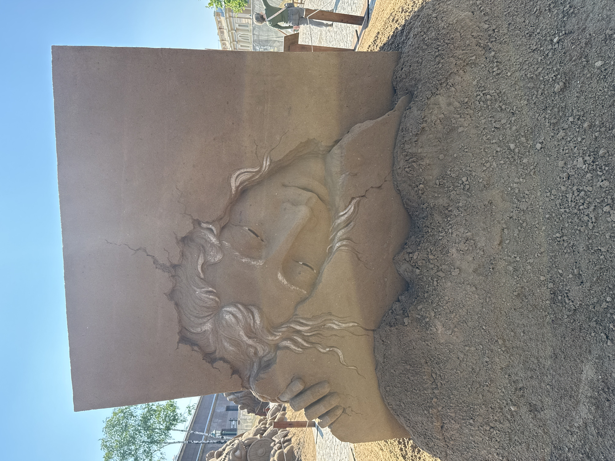 The image shows a close-up of a sand sculpture in Saint Petersburg, Russia. The sculpture depicts a face in profile, with closed eyes and stylized hair. The face appears to be emerging from a rectangular block. The right side of the sculpture is bordered by a pile of uncarved sand. In the background, out of focus, are buildings typical of Saint Petersburg architecture, including windows and facades, and at least one person on a paved surface. The sky is clear and blue.