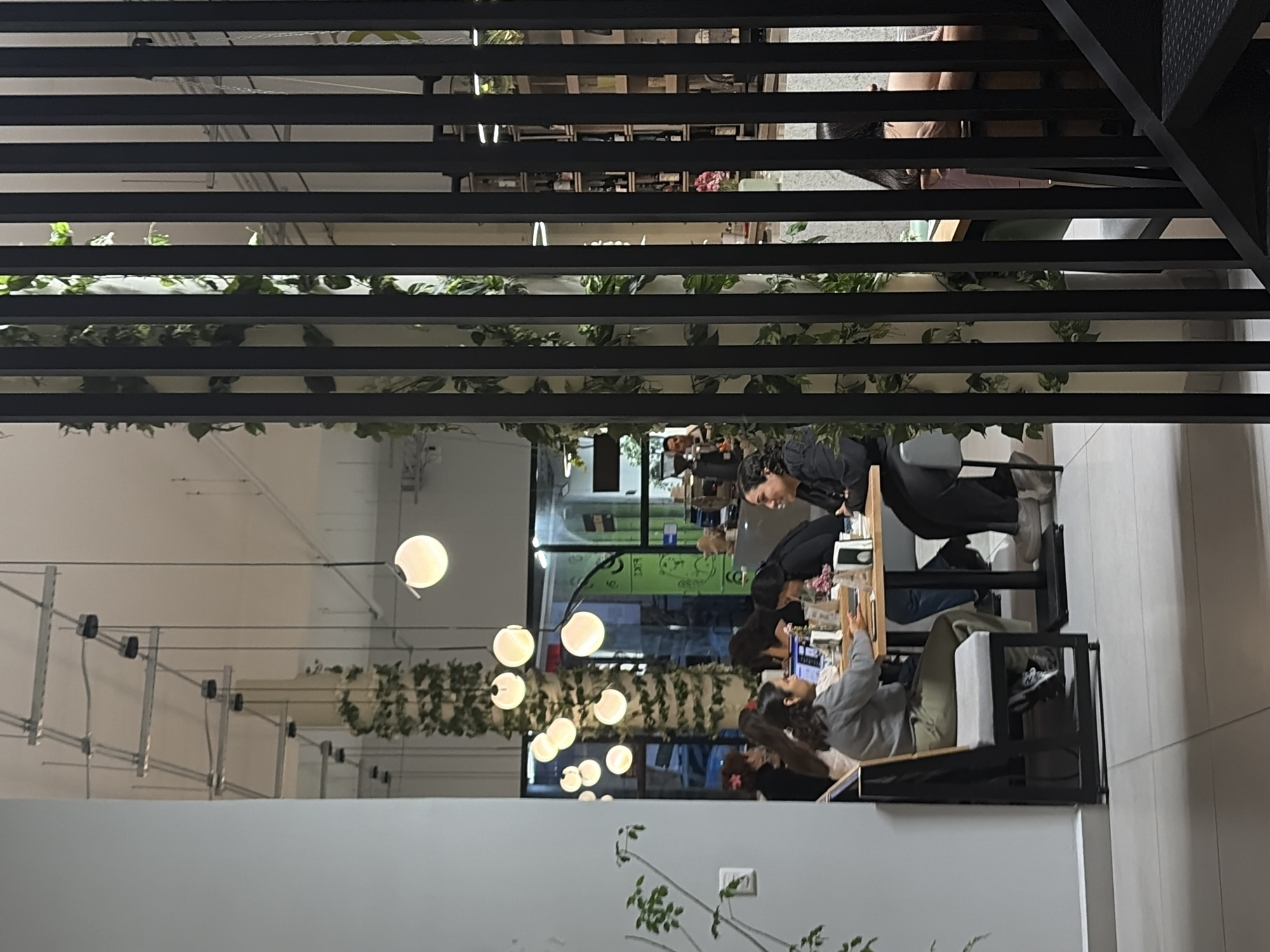 The image depicts an indoor scene in Puebla, Mexico, observed from above, with a perspective through a lattice-like structure. The area is decorated with hanging plants and spherical light fixtures on a branching structure. Several people are seated at tables, some are interacting with phones, and others are engaged in conversation. The background reveals a brightly lit, open space with windows and additional plant arrangements. The setting appears to be a cafe or a communal workspace.