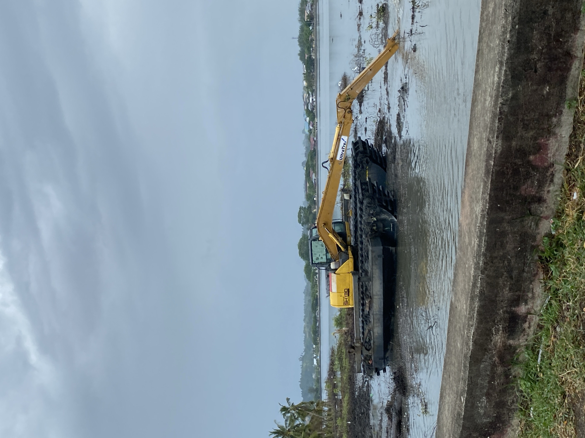 The image, taken in Lhokseumawe, Indonesia, displays a large yellow amphibious excavator situated in a body of water under an overcast sky. The excavator, identified by "SH130" on its side and "UltraTrel" on its boom, appears to be a Komatsu model. Its long arm is extended into the water to the right, indicating it is either performing or about to perform dredging, excavation, or clearing operations in this marshy or flooded environment.
The scene is set in a wetland or waterfront area during daytime, characterized by a muted, grey sky. In the immediate foreground on the right, a weathered concrete barrier or wall with some grass on its edge separates the viewer from the water. The water itself is calm but appears somewhat murky, containing debris and scattered vegetation. Further into the water, towards the background, several small white birds, possibly egrets, are visible standing amidst the aquatic plants.
Across the expanse of water, a distant shoreline with a line of trees and various buildings can be seen, suggesting a populated area. Lush green vegetation, including distinct palm trees in the lower left, reinforces the tropical setting. The overall impression is one of ongoing environmental or infrastructure work in a semi-urban, aquatic landscape.