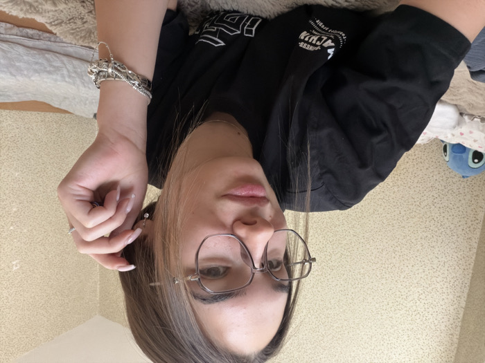 A young woman is depicted in a close-up, chest-up shot in an indoor setting within Sofia, Bulgaria. She has long brown hair with lighter streaks and wears octagonal-framed glasses. Her expression is a slight smirk, and she looks towards the viewer. She is dressed in a black short-sleeved t-shirt with white text and a design partially visible on the front. Her right hand is raised, with fingers near her right ear, displaying manicured nails with light polish. She wears a small, dangling silver-colored earring with a clear stone in her right ear, a silver-toned ring with small stones on her right ring finger, and multiple silver-toned bracelets on her right wrist. She appears to be reclining on a textured, light brown or beige blanket or rug. In the background to her left, a blue stuffed animal resembling Stitch rests on a white pillow with a pink floral pattern. The wall behind her is covered with a light beige, speckled, textured wallpaper. A folded grey textile is visible on a surface to her right.