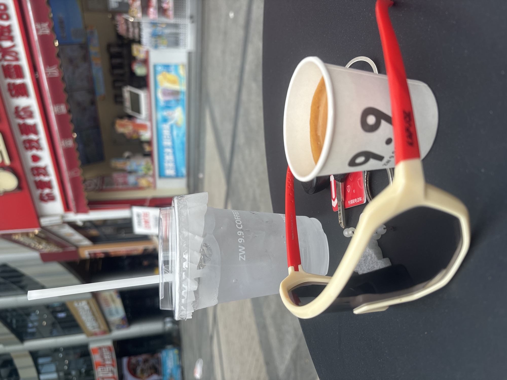 In the Nanhai District of China, on a black table, a pair of beige sunglasses with red arms sits in the foreground. To the right of the sunglasses is a small white paper cup containing a brown beverage, with the number "9.9" printed on it. Beside the cup is a set of keys and a red card. Further back, a clear plastic cup filled with ice and water stands with a white straw. In the background, a red-awninged storefront displays Chinese characters and pictures of drinks, suggesting a beverage shop. The setting appears to be an outdoor seating area.