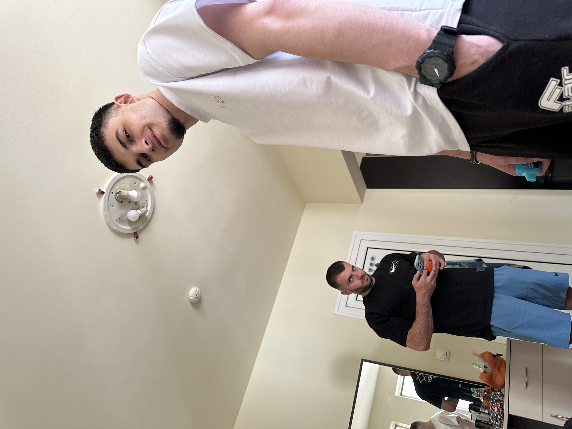 The image depicts two men in an interior setting. The first man is wearing a white t-shirt and black shorts with a wristwatch on his left wrist. He is positioned closer to the camera and appears to be holding a small blue object in his left hand. The second man, seen in the background, is wearing a black t-shirt and light blue shorts, and he is holding a mobile phone in one hand and a small orange object in the other. He is standing in front of a white door frame. The room includes a light fixture on the ceiling and a mirror reflecting both men. There are no discernible elements that definitively place the location in the city of Sofia, Bulgaria.