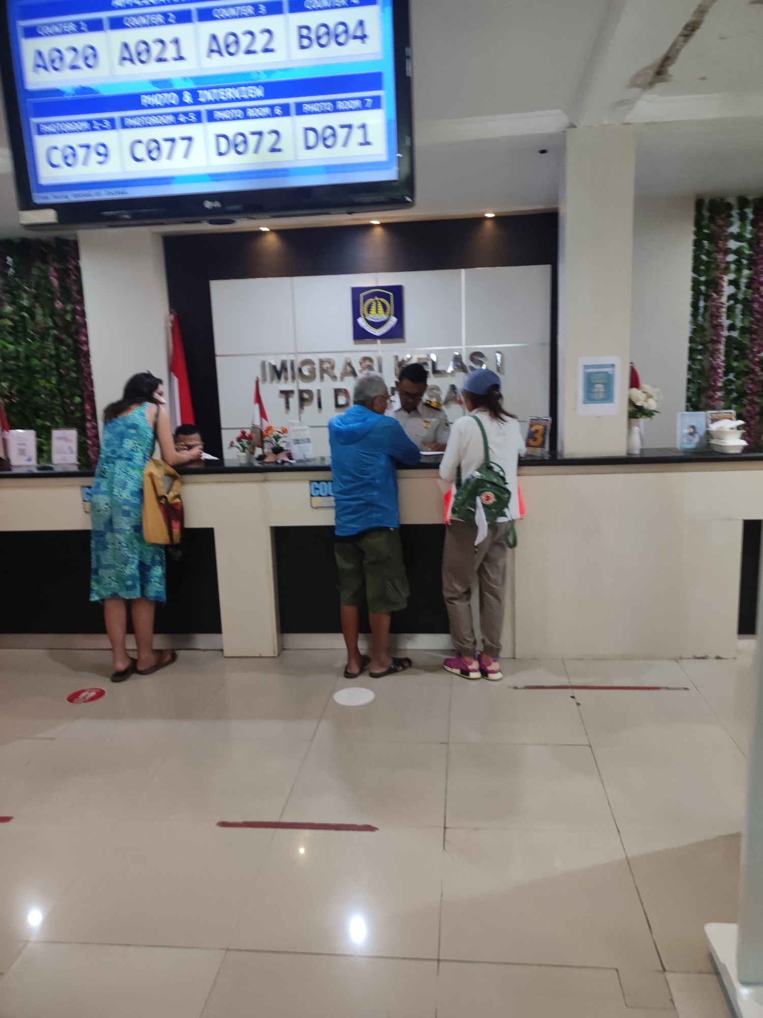 The image depicts the interior of the Immigration Office (IMIGRASI TPI) in Denpasar, Indonesia. There are several people standing in front of a counter, presumably waiting to be served. The counter is manned by an officer in uniform. A digital display above the counter shows a queue system with numbers such as "A020," "A021," "A022," "B004," "C079," "C077," "D072," and "D071." The floor has markings indicating social distancing measures. The scene appears to be a typical day at the immigration office.