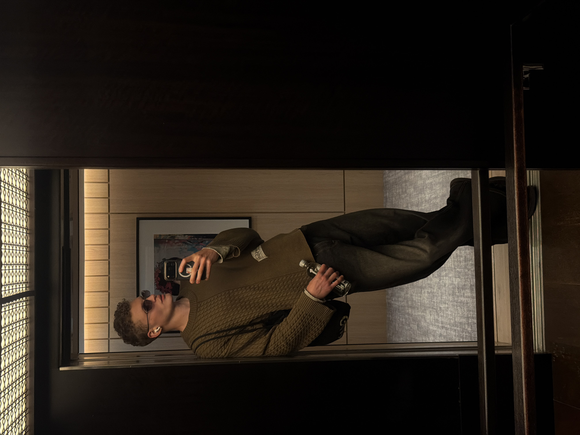A young man is reflected in a mirror, holding a phone in one hand and a small bottle in the other. He has short, curly hair and is wearing sunglasses and an earbud. He's dressed in a patterned sweater and dark pants. The background includes decorative window panes, wooden panels, and a framed picture. The image appears to be indoors, though the exact location in London, United Kingdom, cannot be determined solely from the visual information provided.