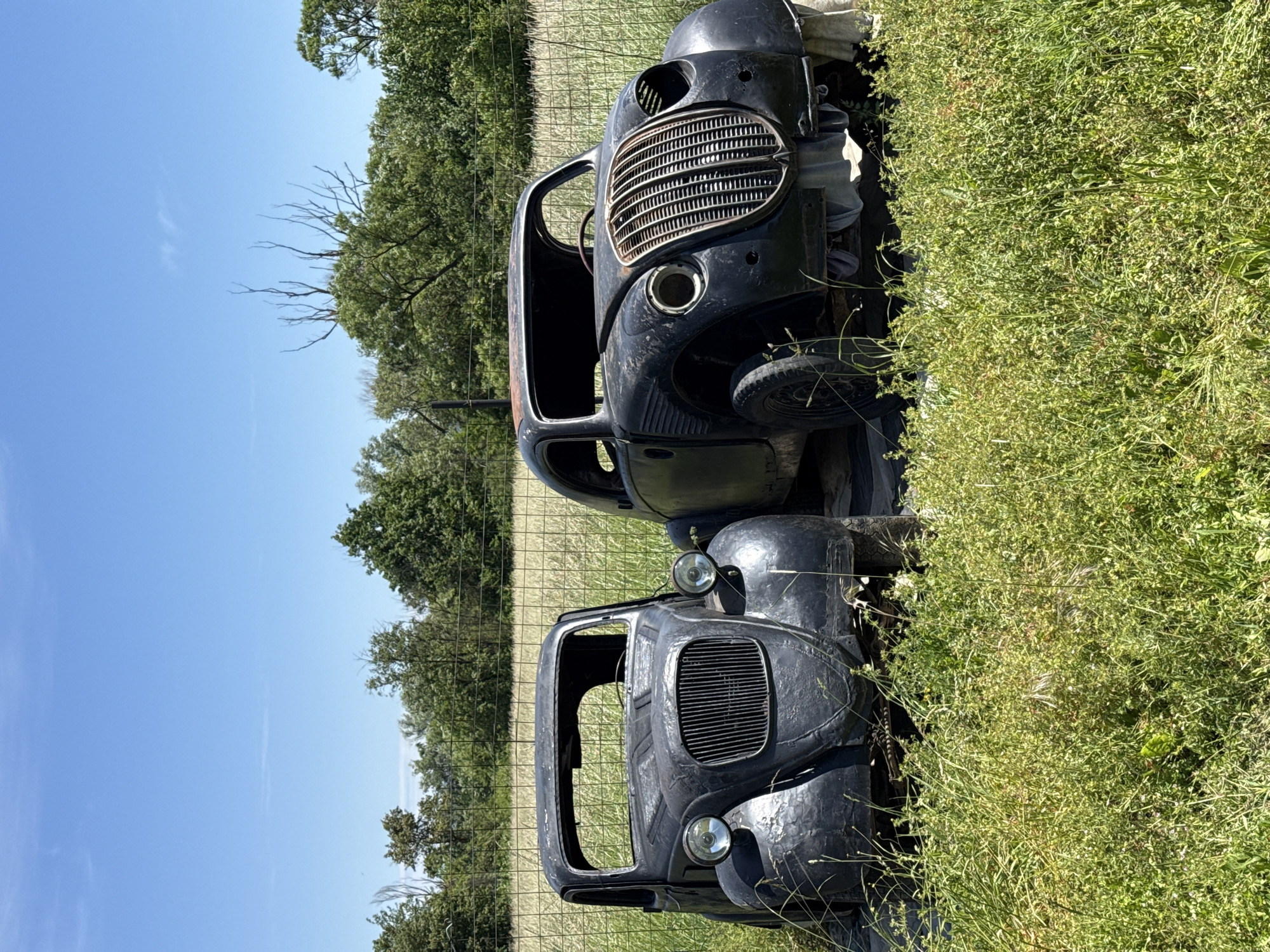 Two black, dilapidated vintage car bodies stand outdoors, in a field of green grass and vegetation. The lower car has two intact headlights and a vertical-slatted grill. The upper car is more deteriorated, missing headlights, and only has partial tires. A wire fence is visible behind the cars, with trees and a clear blue sky in the background. The location is Omurtag, Bulgaria.