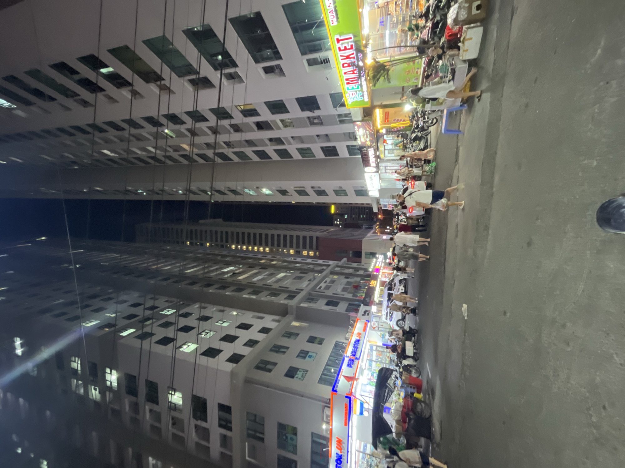 The image shows a street scene at night in Bac Nha Trang Ward, Vietnam. High-rise apartment buildings line one side of the street, with commercial establishments at street level. Pedestrians are walking along the sidewalk, and parked motorbikes are visible. Street vendors or shops are present, indicated by lights and displayed goods. The street is paved and appears to be well-lit. The lighting suggests it is evening or nighttime.
