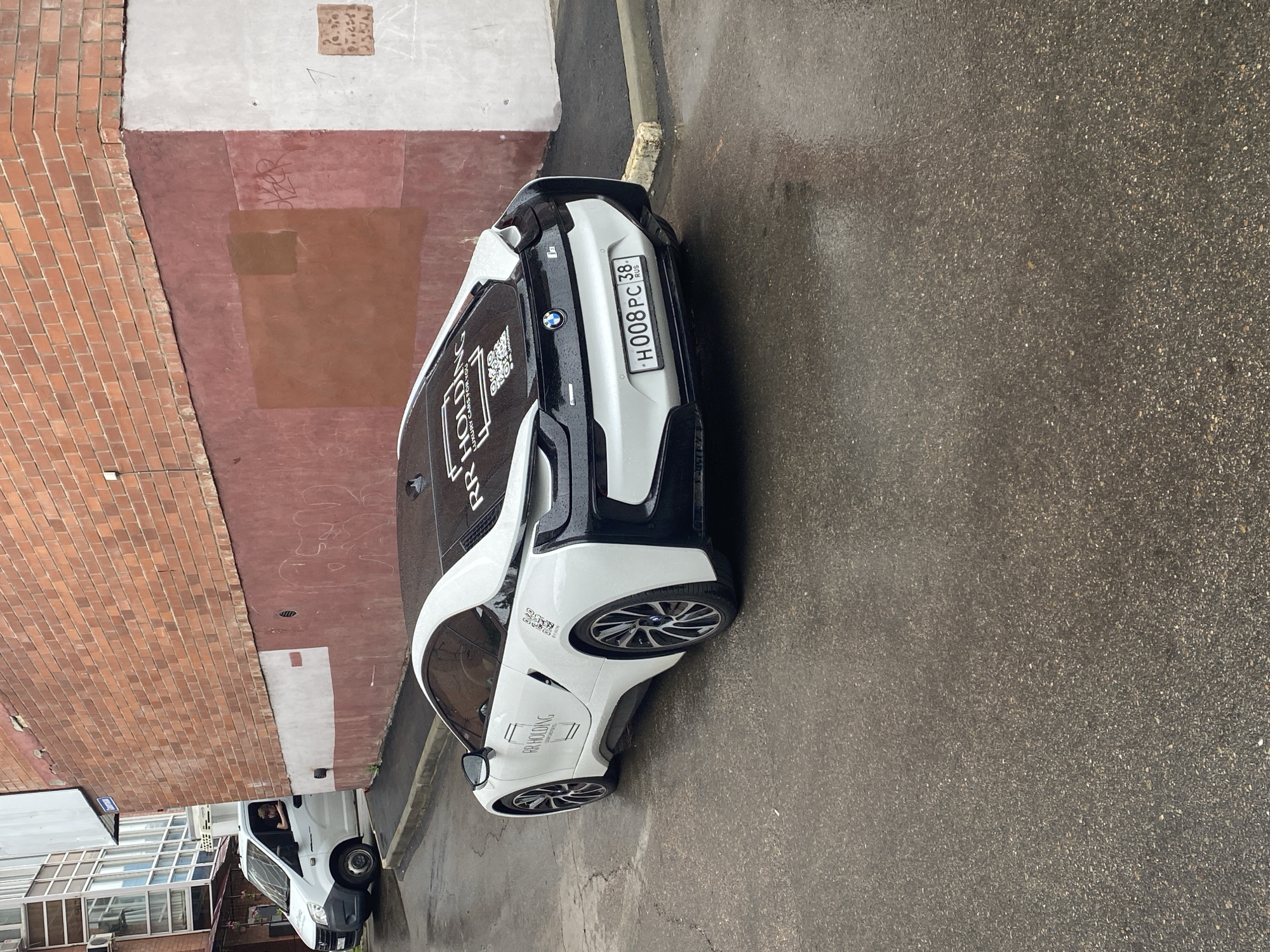 A white and black BMW i8 is parked on a wet asphalt street in front of a brick building. The car has "RR HOLDING" and "LUXURY CARS FOR YOU" decals on its rear window and a QR code. The license plate reads "H008PC 38 RUS". A white van with a person inside is parked behind the BMW. The building has a red-painted lower section and a light-colored upper section. The ground is wet, indicating recent rain.