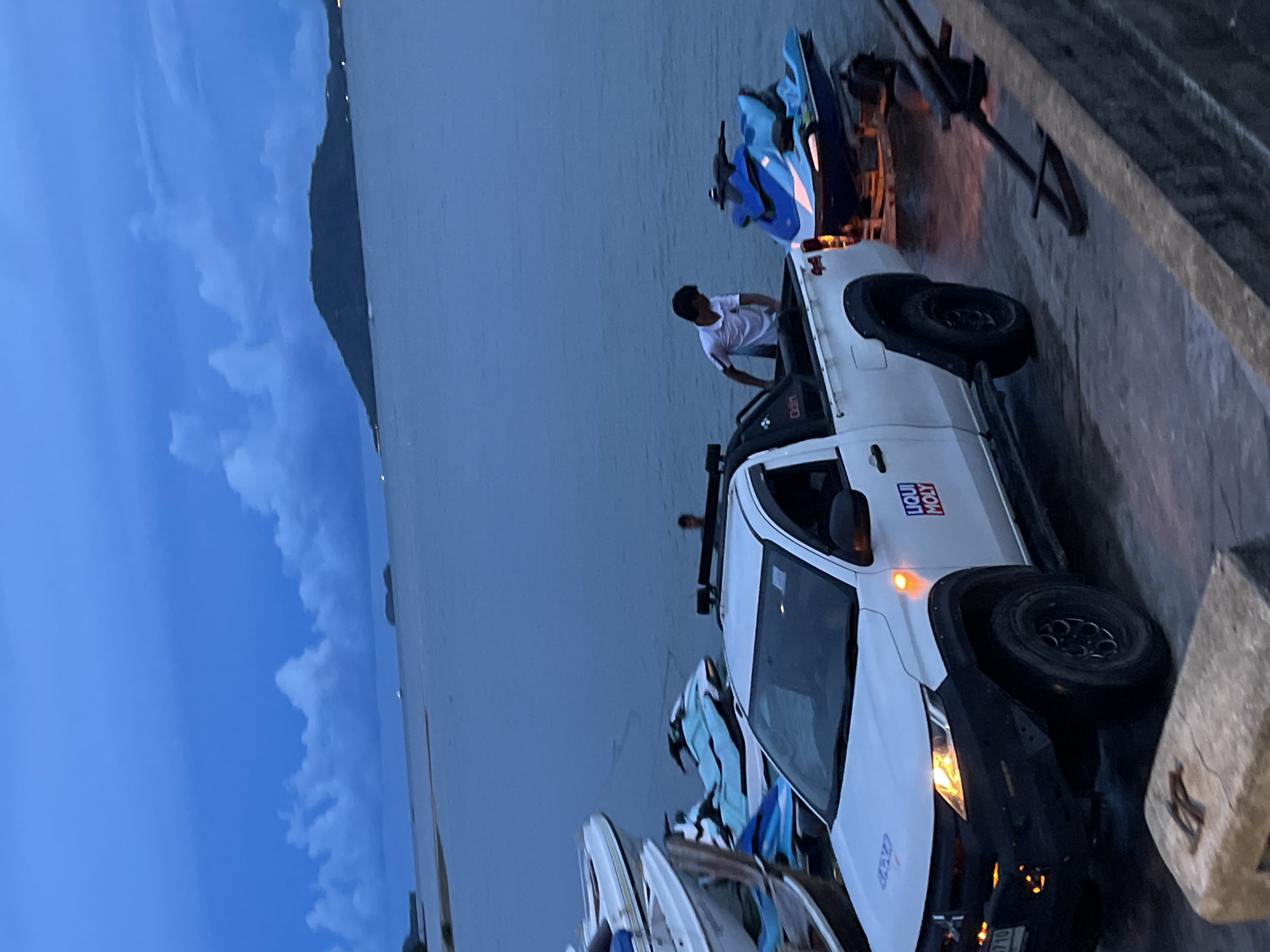 A man in a white t-shirt is leaning into the bed of a white pickup truck, which is parked on what appears to be a ramp or dock. The truck is loaded with what look like jet skis or personal watercraft. The scene is set at dusk, with a twilight sky and the ocean in the background. The water is calm and a dark, deep blue, reflecting the sky. A distant landmass, possibly an island or mountain range, is visible on the horizon. The overall mood is peaceful and serene, with a hint of activity as the man attends to the cargo. Visible text on the truck includes a "LIQUI MOLY" sticker.