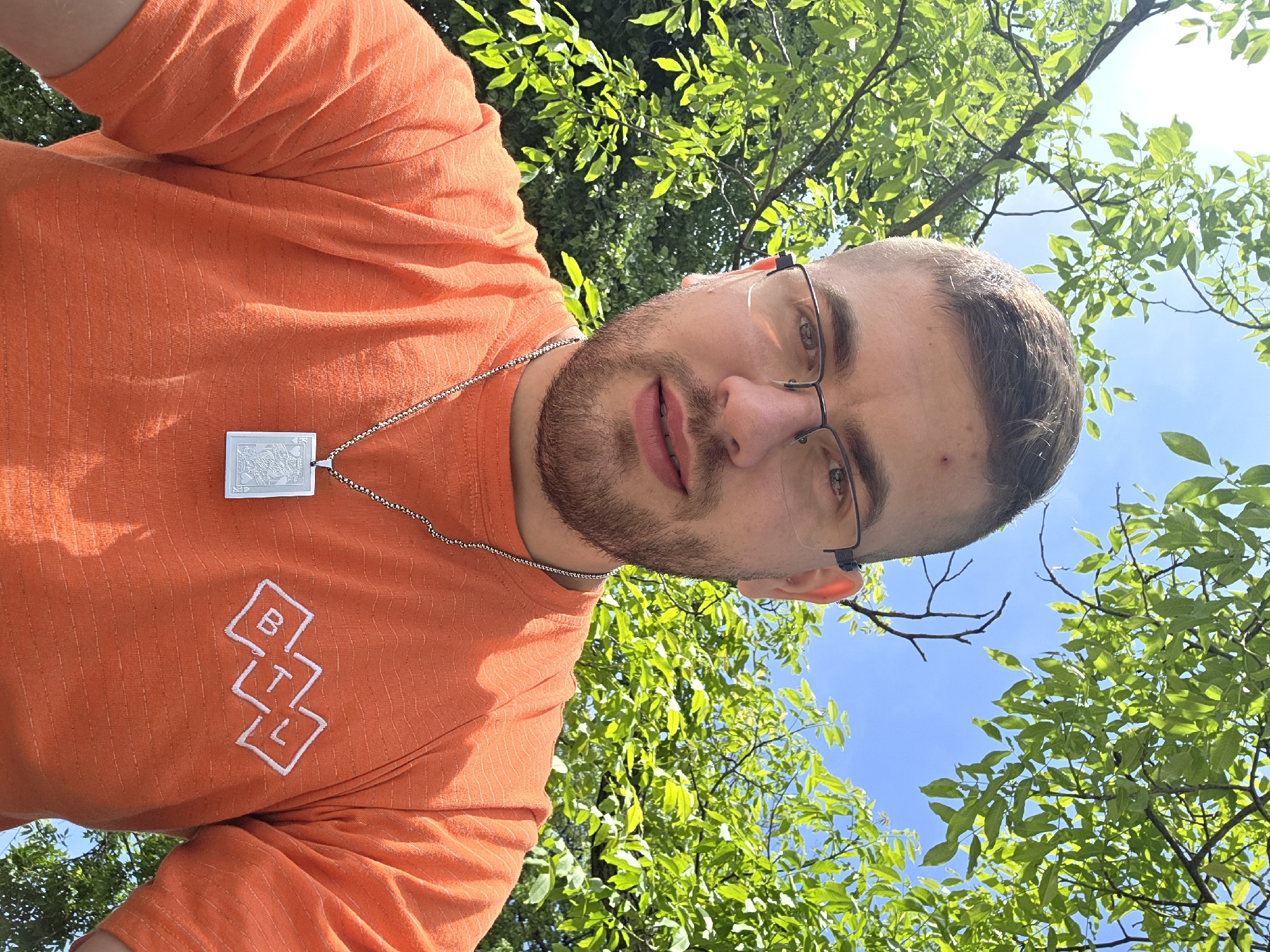 A man with short hair and a beard, wearing glasses and an orange t-shirt with the letters "BTL" printed on it, is shown in a selfie. He is wearing a silver necklace with a rectangular pendant. The background consists of green leaves and branches of trees against a blue sky. The location in the city of Plovdiv, Bulgaria is not identifiable from the image.