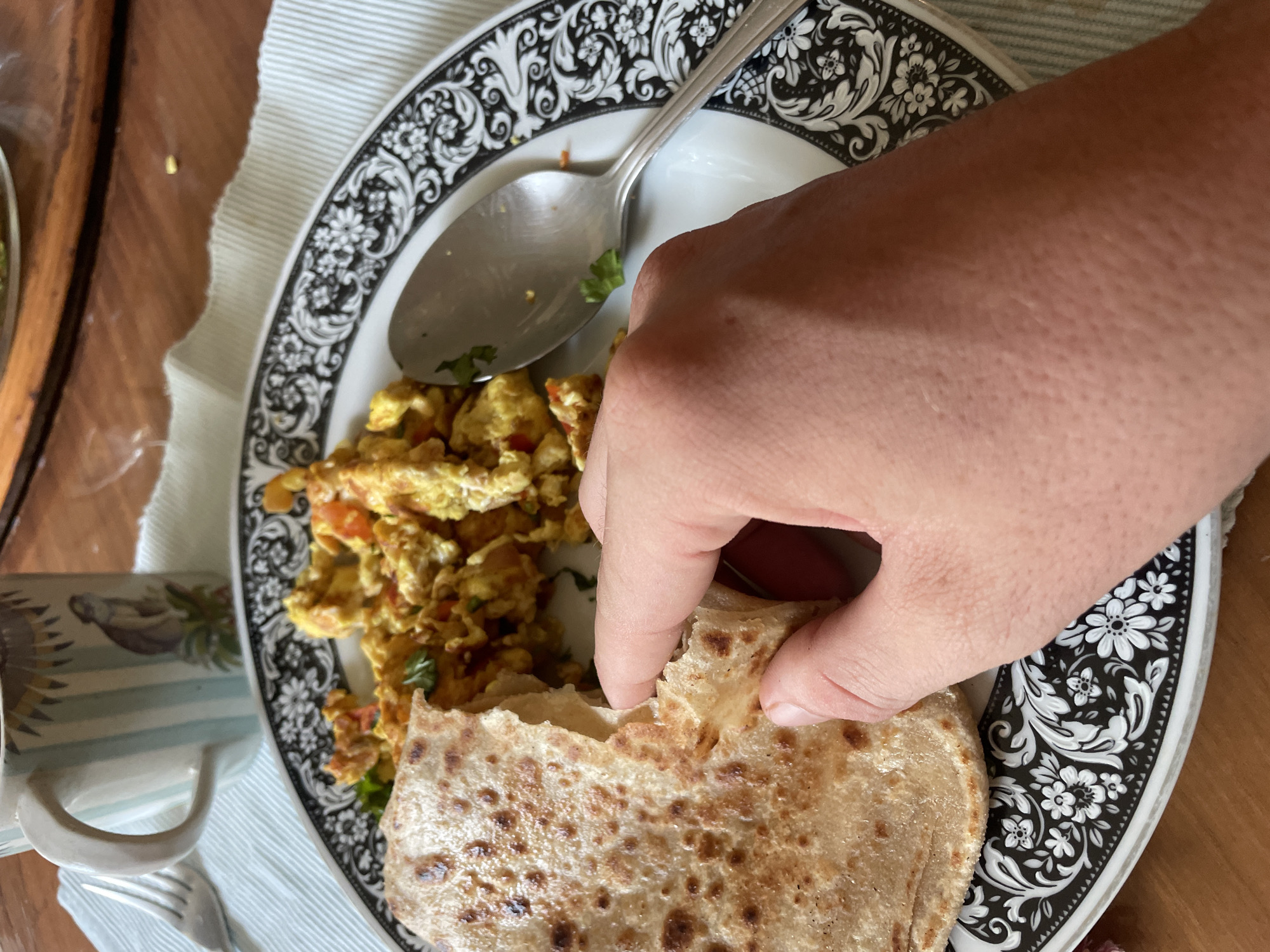 A first-person perspective image shows a plate with a portion of scrambled eggs and a flatbread being torn apart by a human hand. A silver spoon is on the plate next to the eggs. The plate has a black and white floral pattern around the edge. A patterned mug and silverware are visible in the lower-left corner of the image. The table surface is wood.